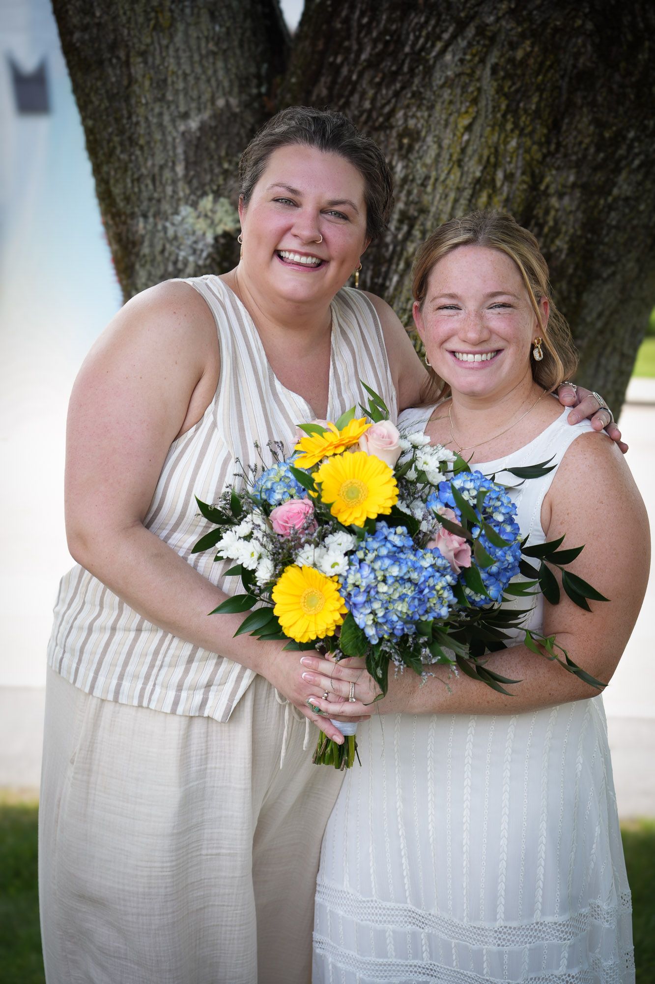 Two women smile together outdoors, sharing a colorful bouquet of yellow, blue, and pink flowers.