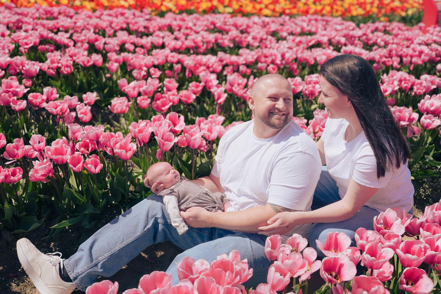 A couple sits relaxed in a lush pink tulip field, sharing a smile while holding their newborn.