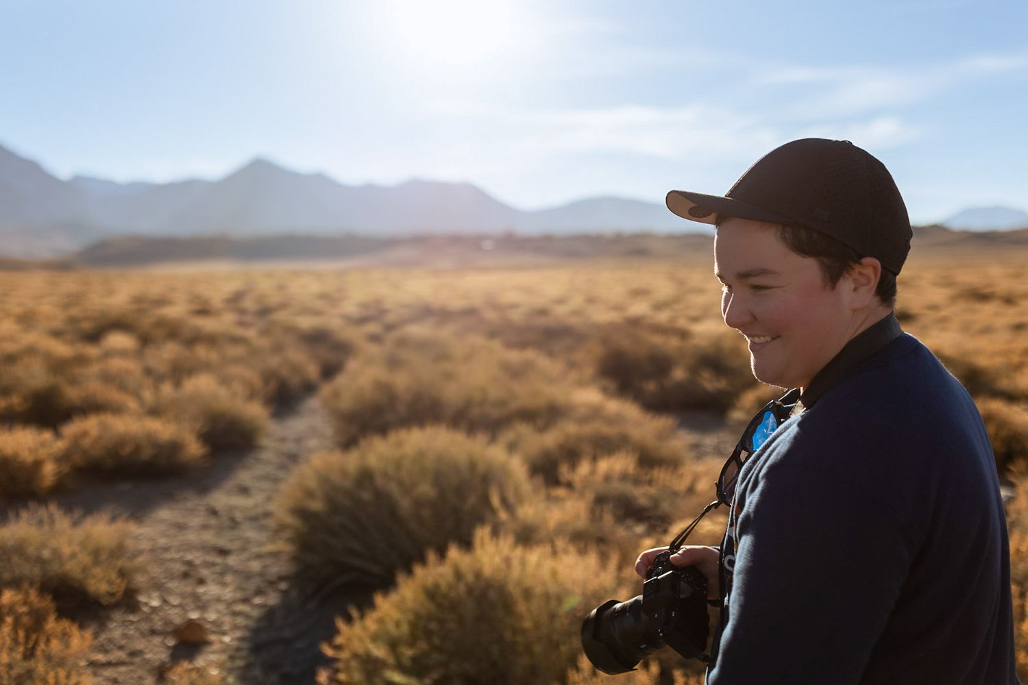Young photographer smiling while holding camera in golden desert landscape with mountains behind.