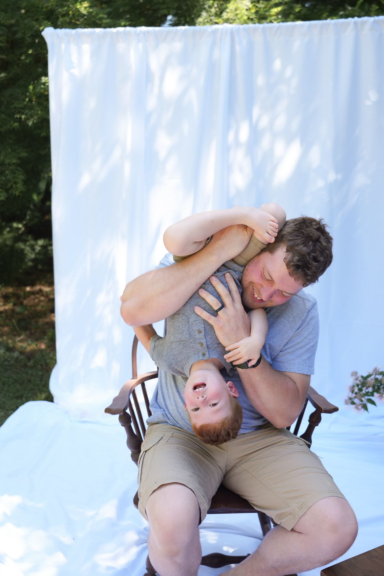 Playful father holding young red-haired son upside down on chair outdoors laughing.
