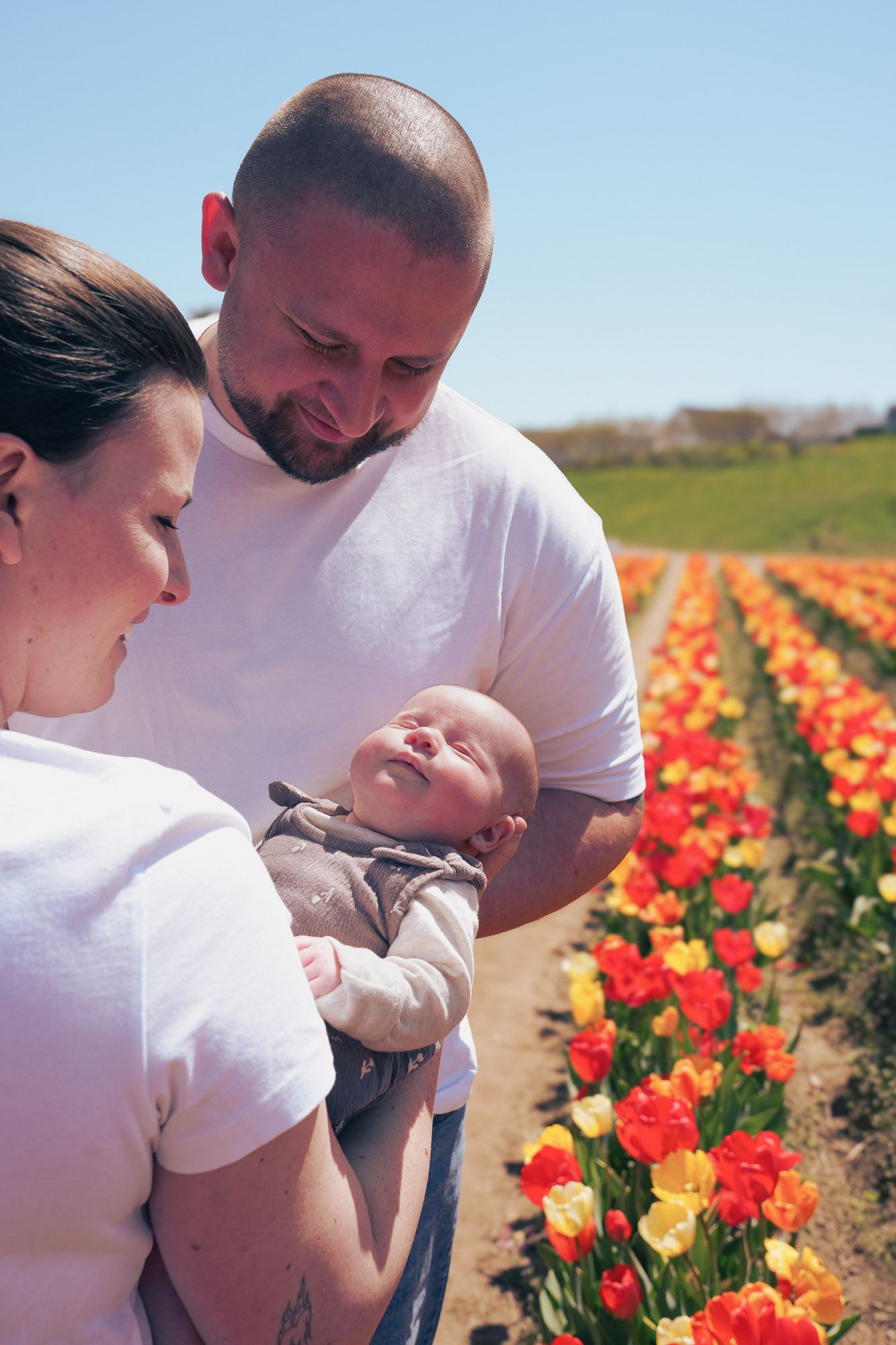 Parents in white shirts lovingly gaze at their newborn baby in a vibrant tulip field on a sunny day.