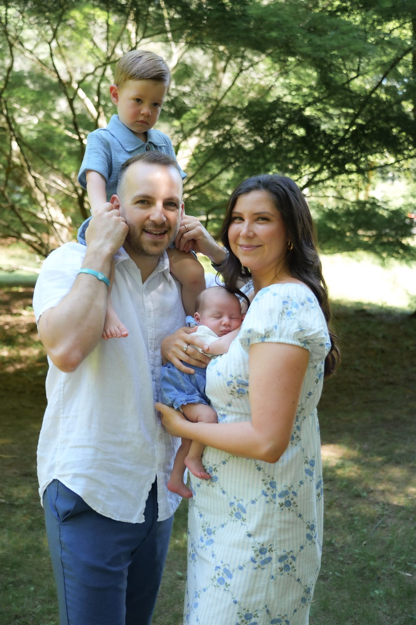 Smiling family portrait outdoors with toddler on dad's shoulders and newborn in mom's arms.