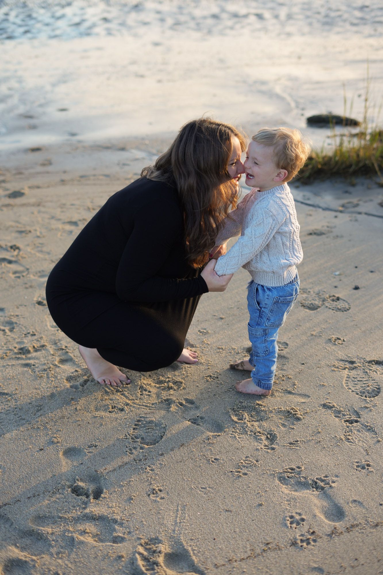 Pregnant woman in black dress kneeling to kiss toddler boy on sandy beach.