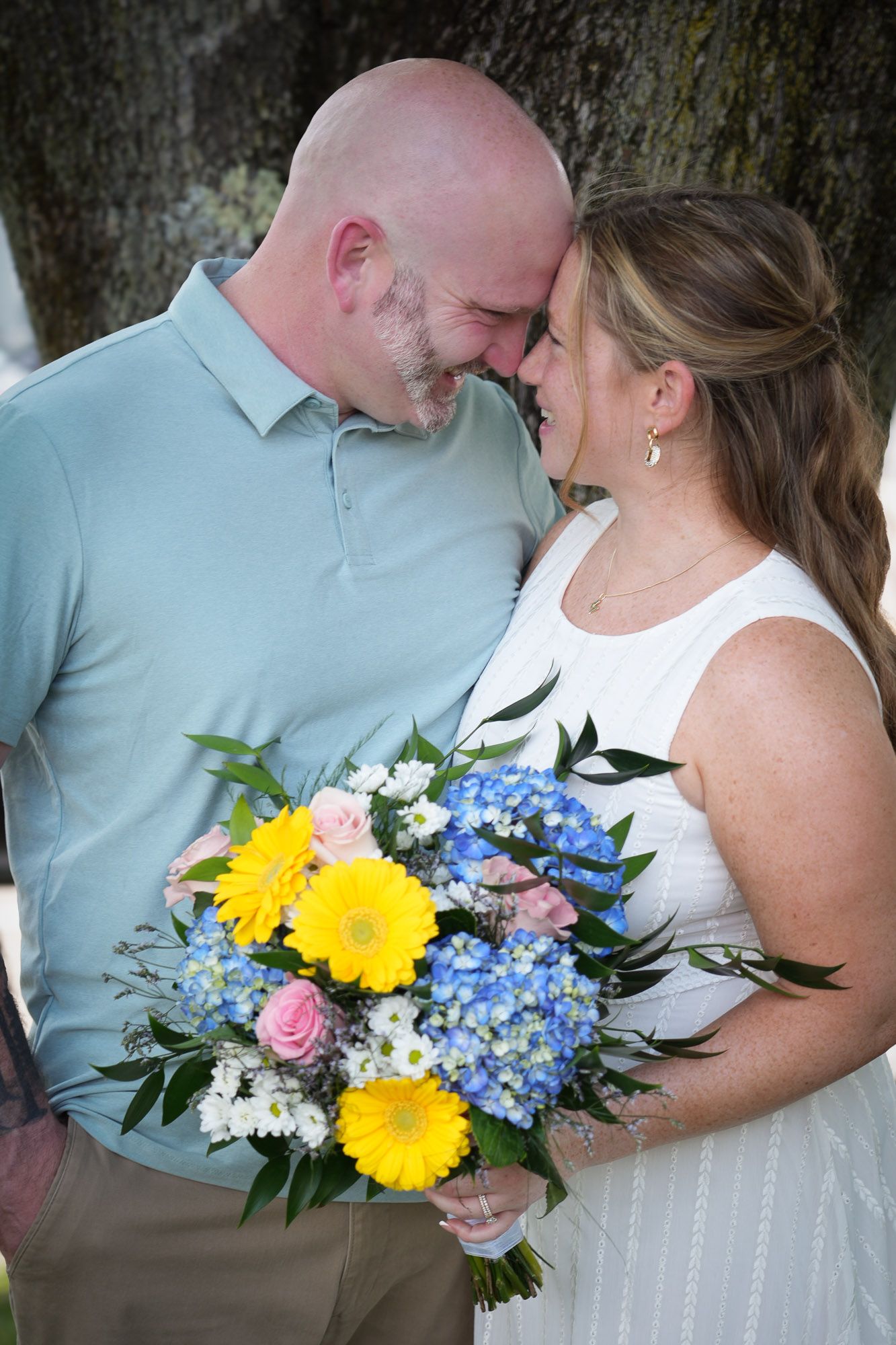 Romantic newlyweds touching foreheads tenderly while holding colorful wedding bouquet with blue hydrangeas.