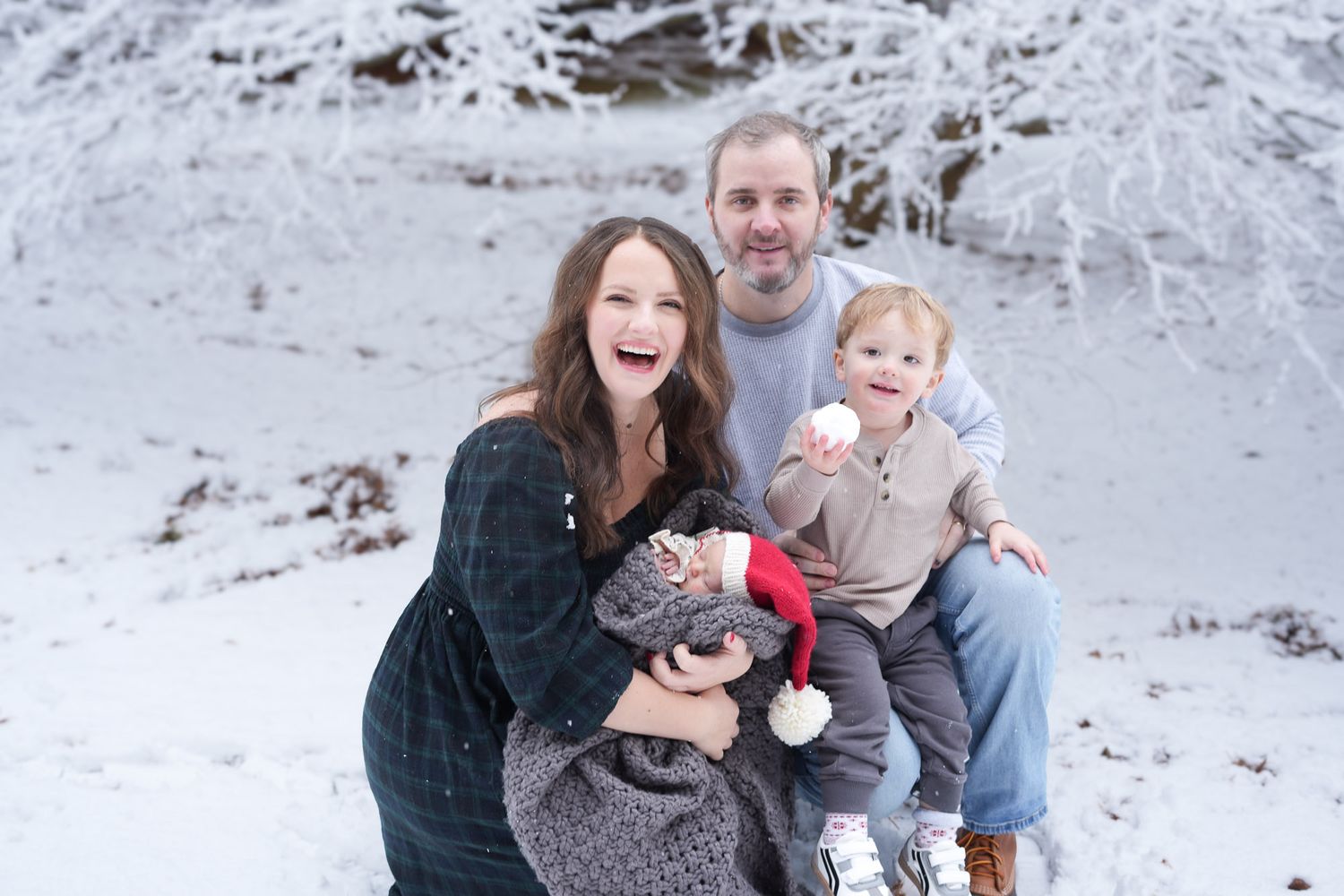 Happy family of four posing together in a snowy winter landscape, parents with toddler and newborn.