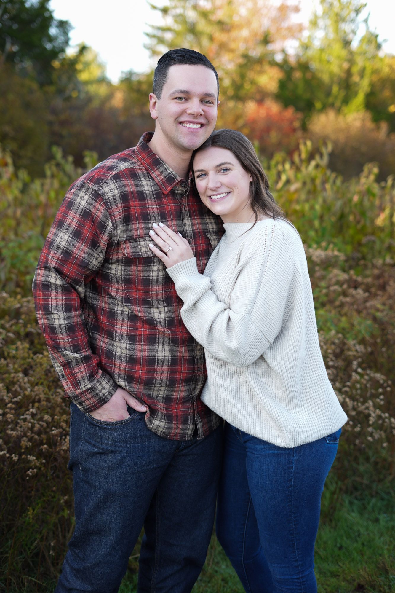 Smiling engaged couple posing together in front of colorful autumn bushes, woman showing engagement ring.