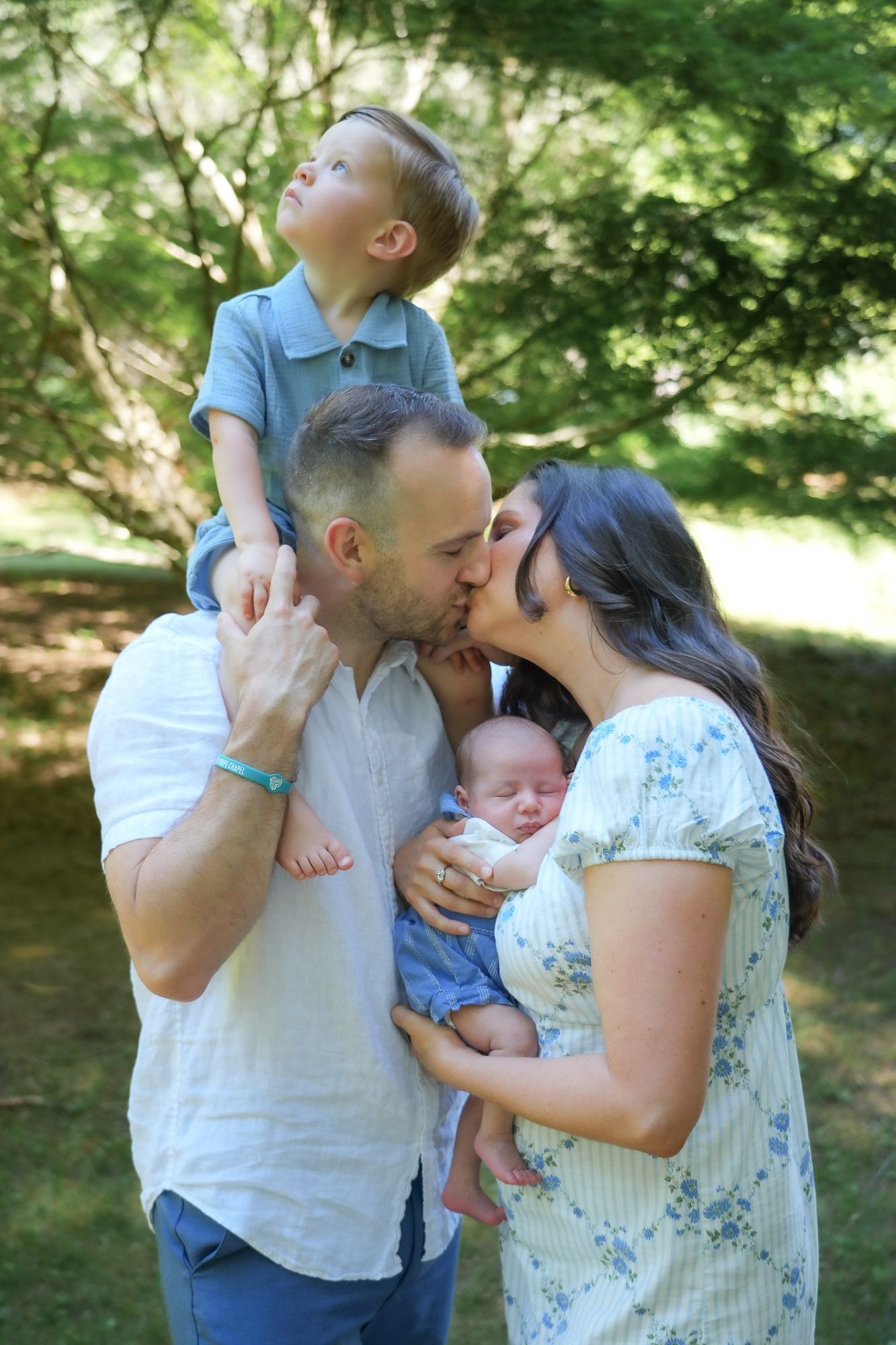 Parents kissing outdoors while father holds toddler on shoulders and newborn baby in arms.