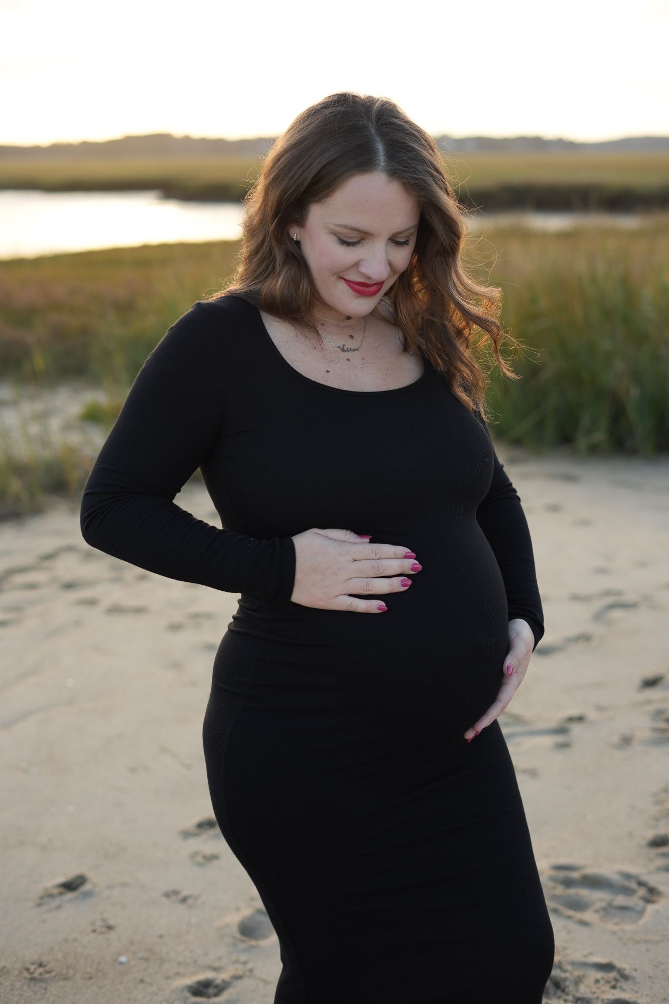 Pregnant woman in black dress looking down at baby bump on beach at sunset.