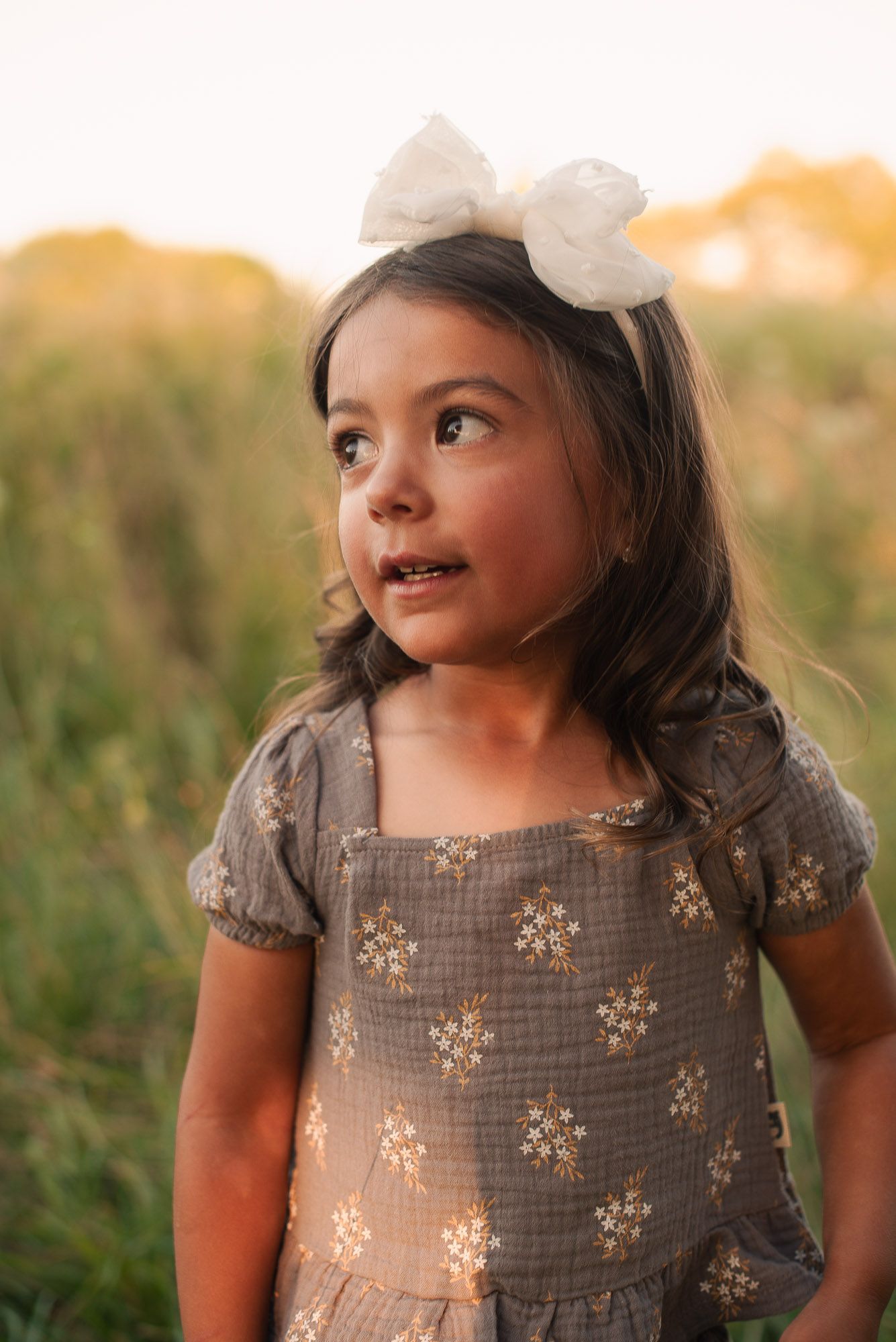 Adorable little girl in a floral dress and white bow headband stands in a sunlit field.