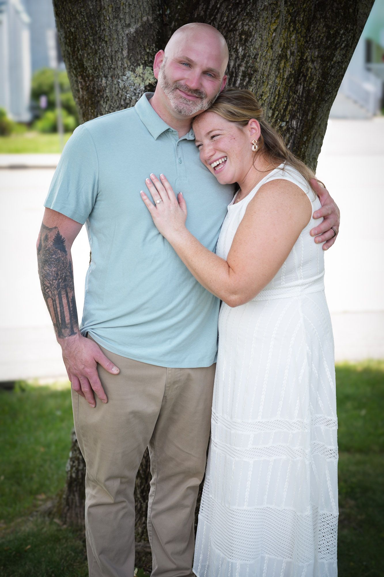 Smiling newlywed couple embracing by a tree, bride in white dress showing engagement ring, groom in blue polo.