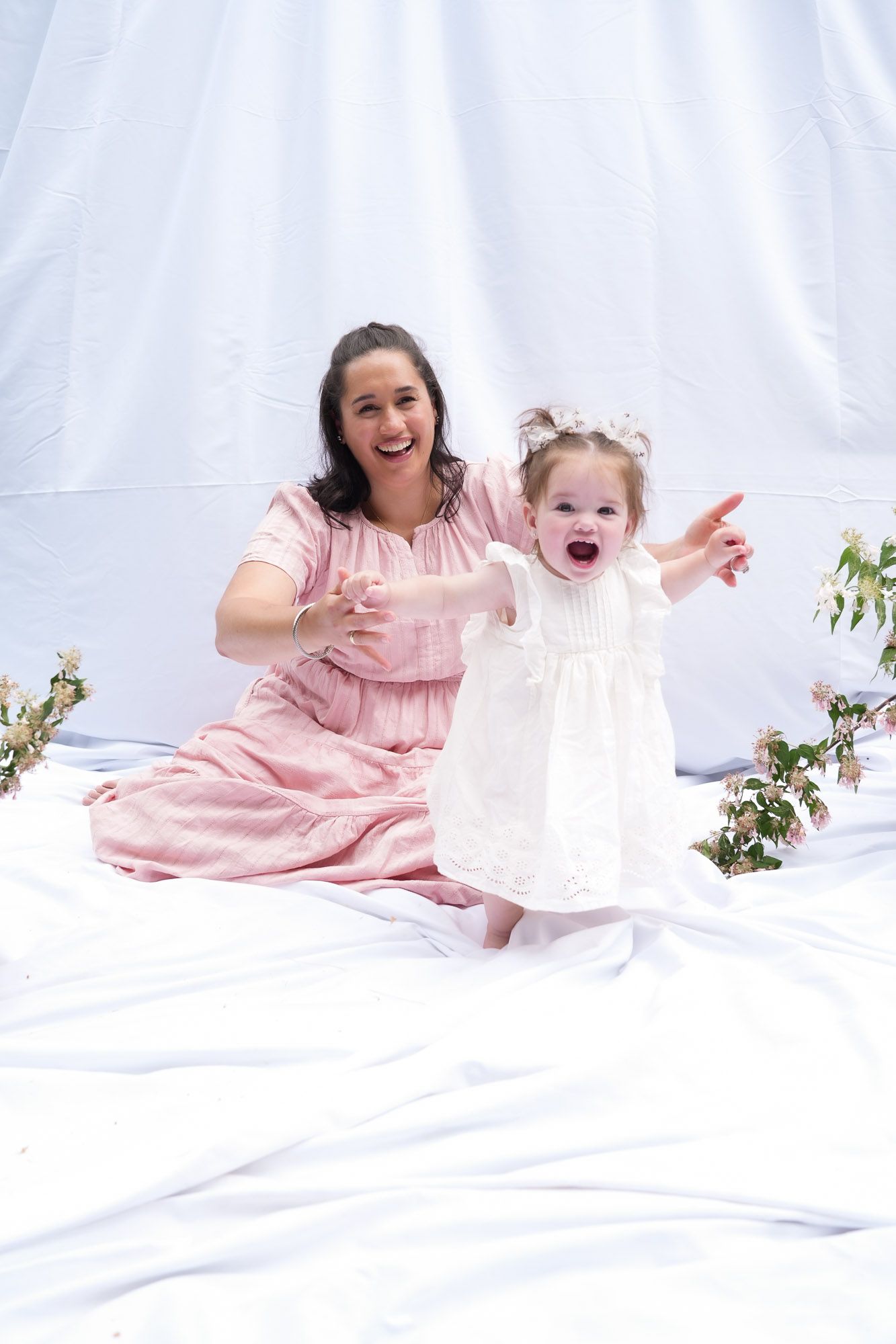 A joyful mother in pink and her laughing toddler daughter in white enjoy a playful moment during an outdoor photo shoot.