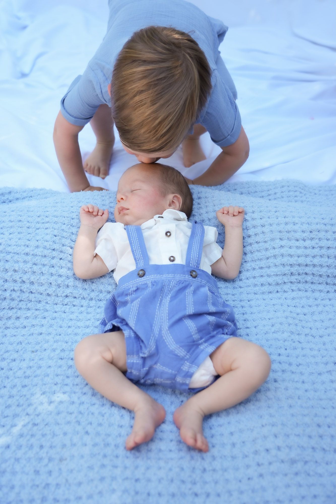 Older child gently kissing a sleeping newborn baby dressed in blue overalls on a soft blanket.