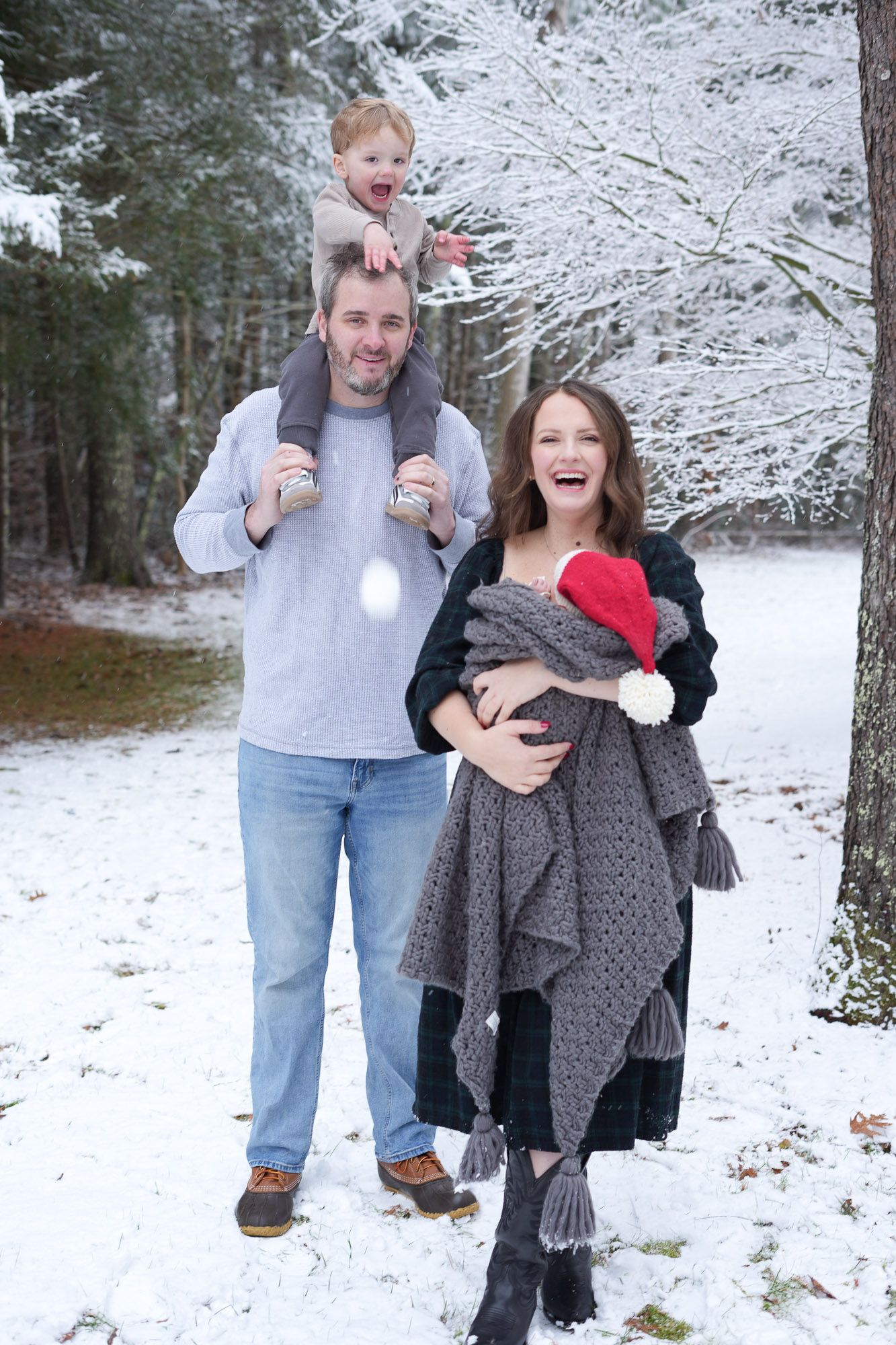 A laughing family of four enjoys a fun winter photo session in the snow, with the toddler perched on dads shoulders.
