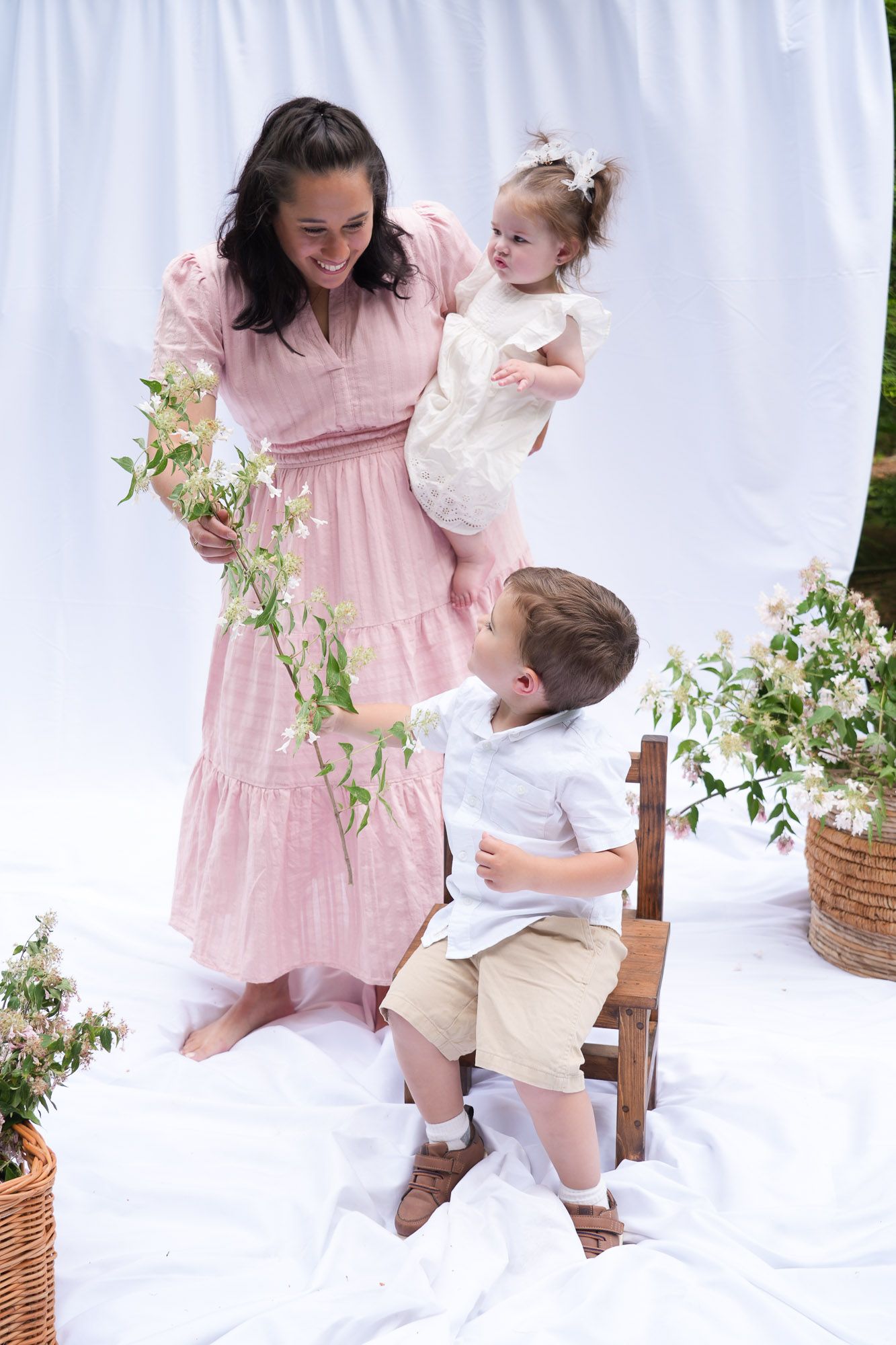 A joyful mother in a pink dress holds her baby girl while her young son sits on a chair among flowers.