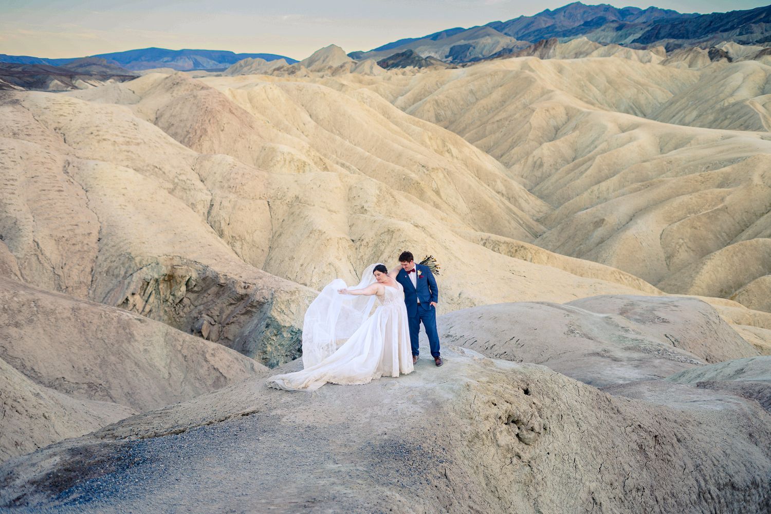 Bride in flowing white gown and groom in blue suit standing amid stunning Death Valley desert landscape.