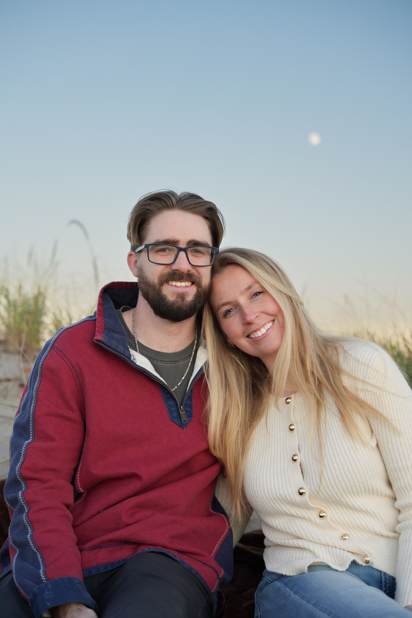 A happy couple posing together on a sandy beach at dusk with the moon above.