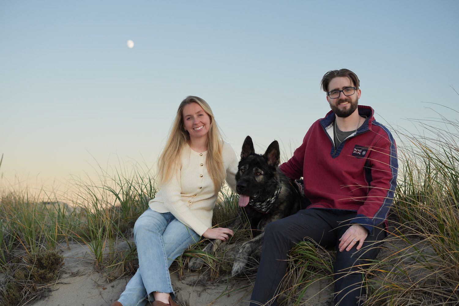 A smiling couple sitting in sand dunes with their German Shepherd dog at sunset.