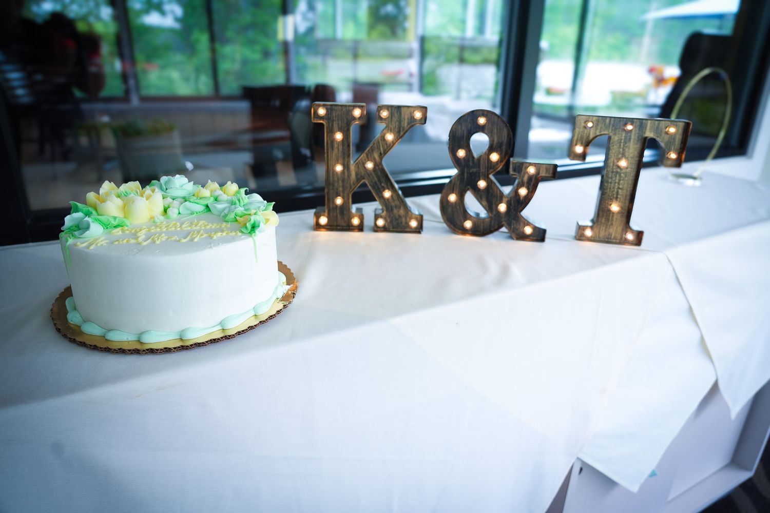 White wedding cake with green floral decorations beside illuminated K&T marquee letter signs on table.