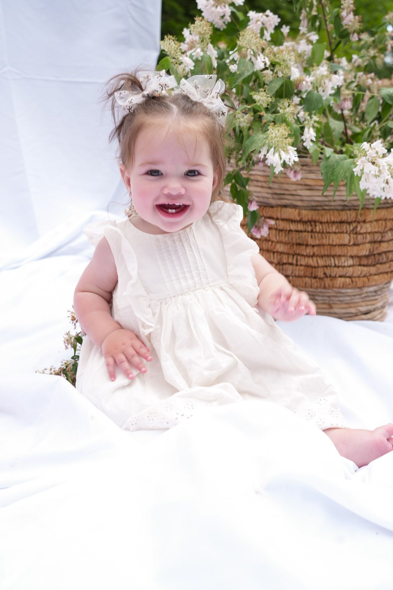 An adorable smiling baby girl in a white dress sits on white fabric next to a wicker basket of flowers.