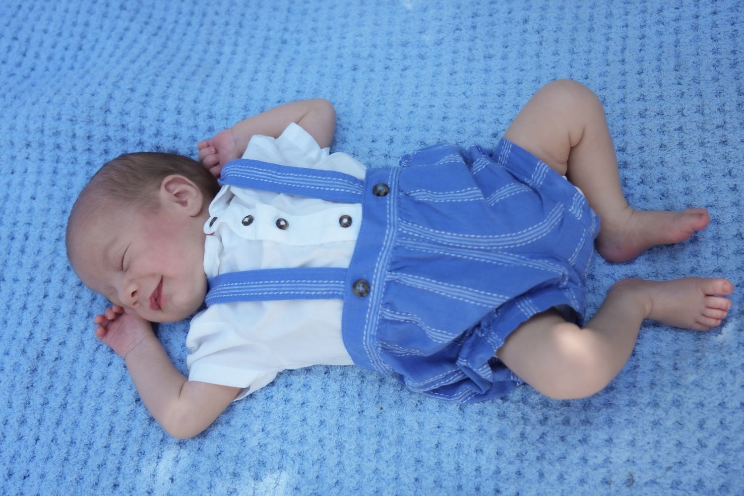 Crying newborn baby boy in blue suspender romper lying on blue knit blanket.