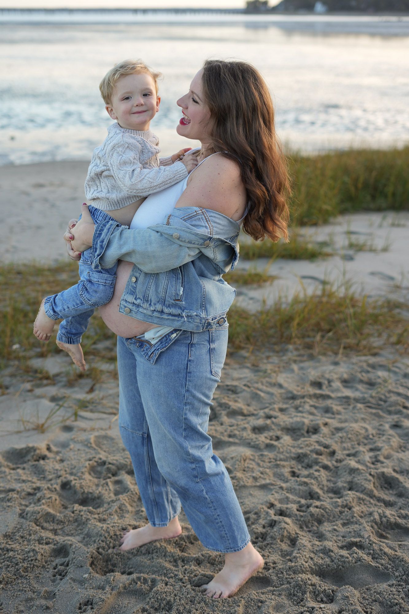 Barefoot pregnant woman in denim holding young child on sandy beach at sunset.
