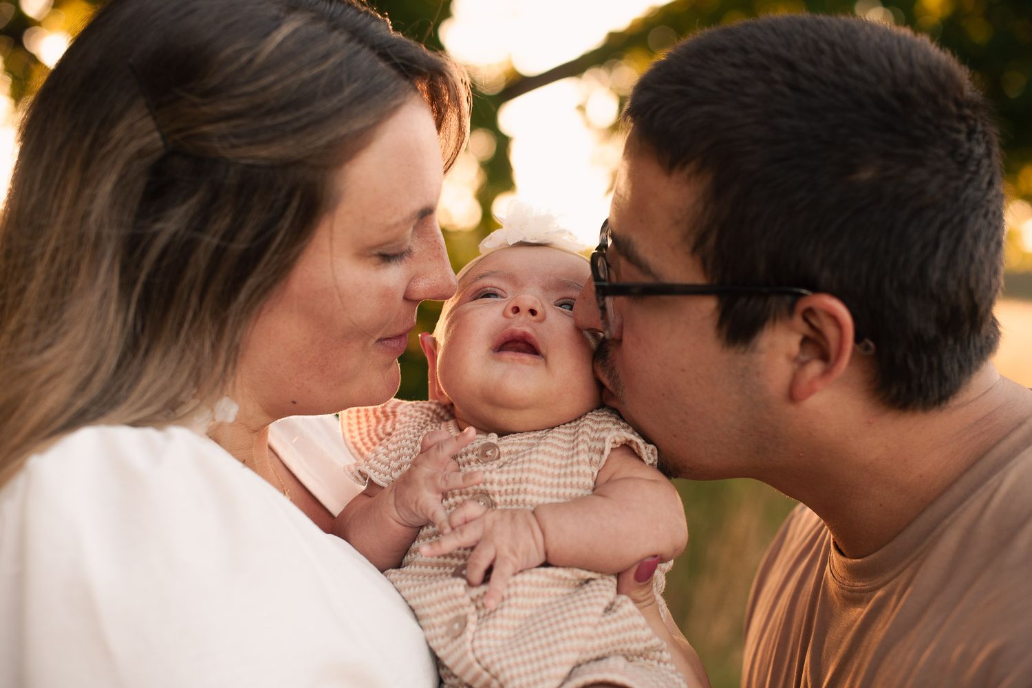 Loving parents share a tender moment kissing their smiling newborn baby on the cheeks outdoors.