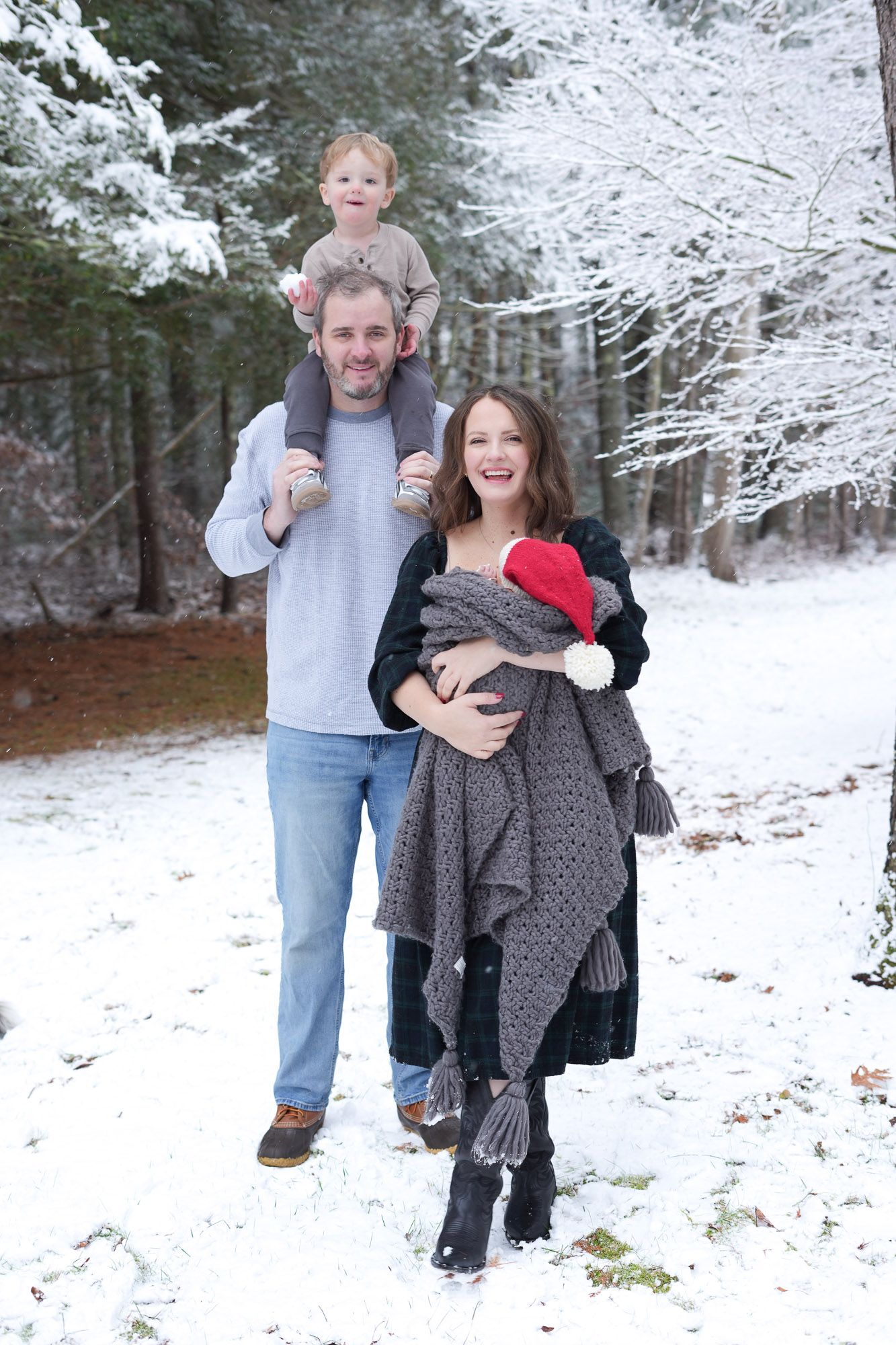 A happy family poses in a snowy forest, with the father carrying their toddler on his shoulders while the mother holds their newborn.