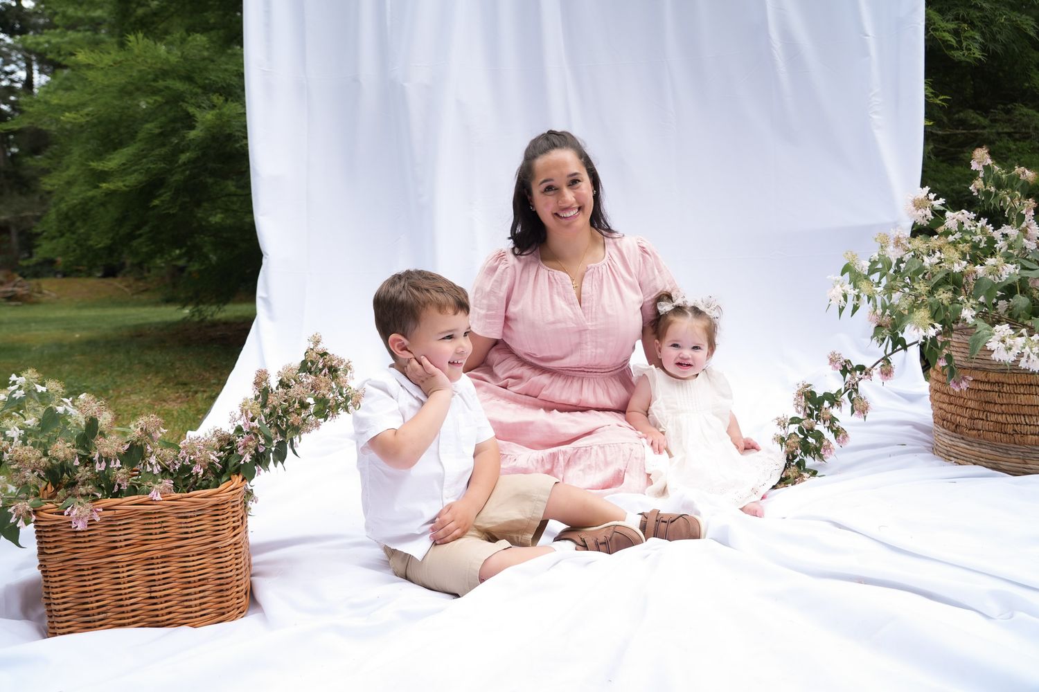 A brunette mother in pink sits with her young son and toddler daughter amid floral arrangements outdoors.