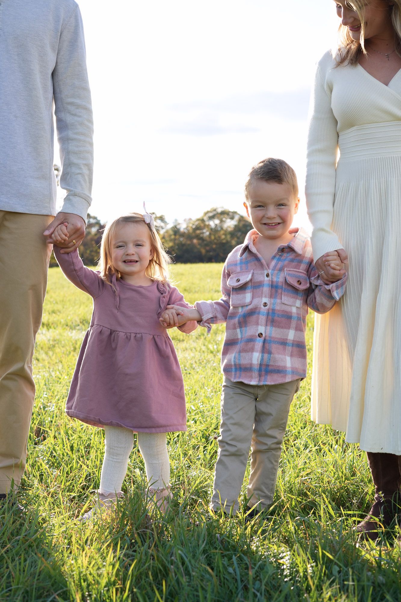 Two smiling young children holding parents hands in a bright green field on a sunny autumn day.