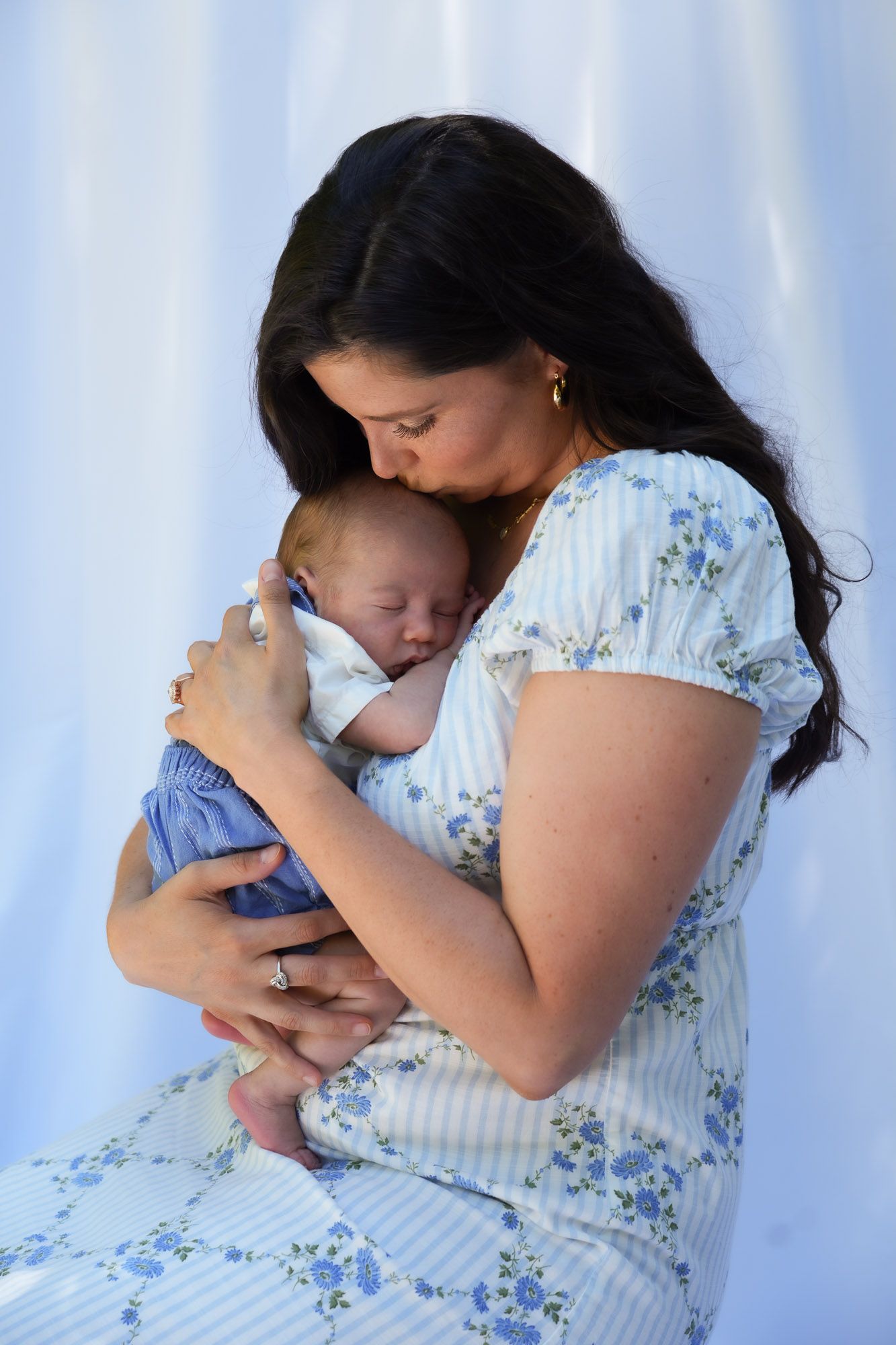 Tender moment of dark-haired mother in floral dress kissing sleeping newborn baby.