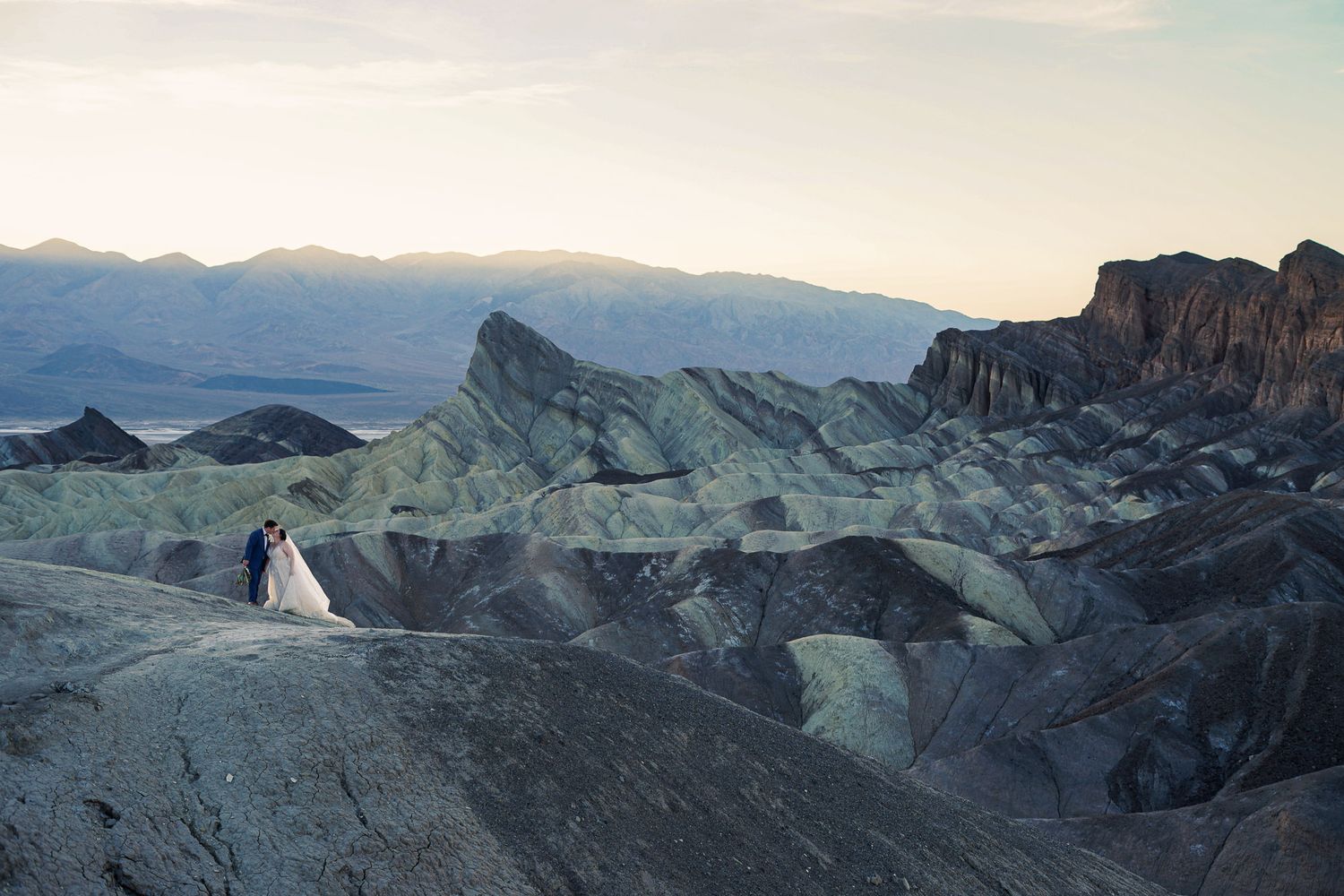 Wedding couple poses atop Zabriskie Point overlooking stunning layered badlands and distant mountains.