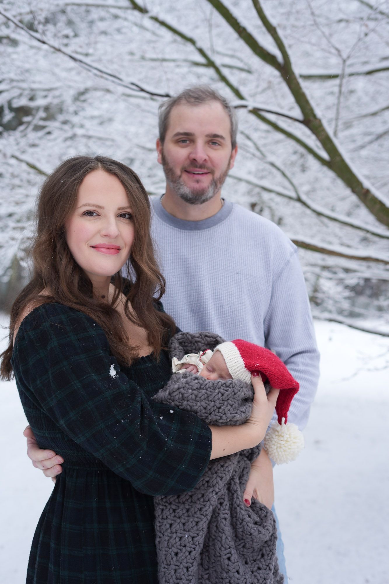 A couple stands in a snowy winter forest, smiling while holding their newborn baby wrapped in a gray blanket with a red Santa hat.