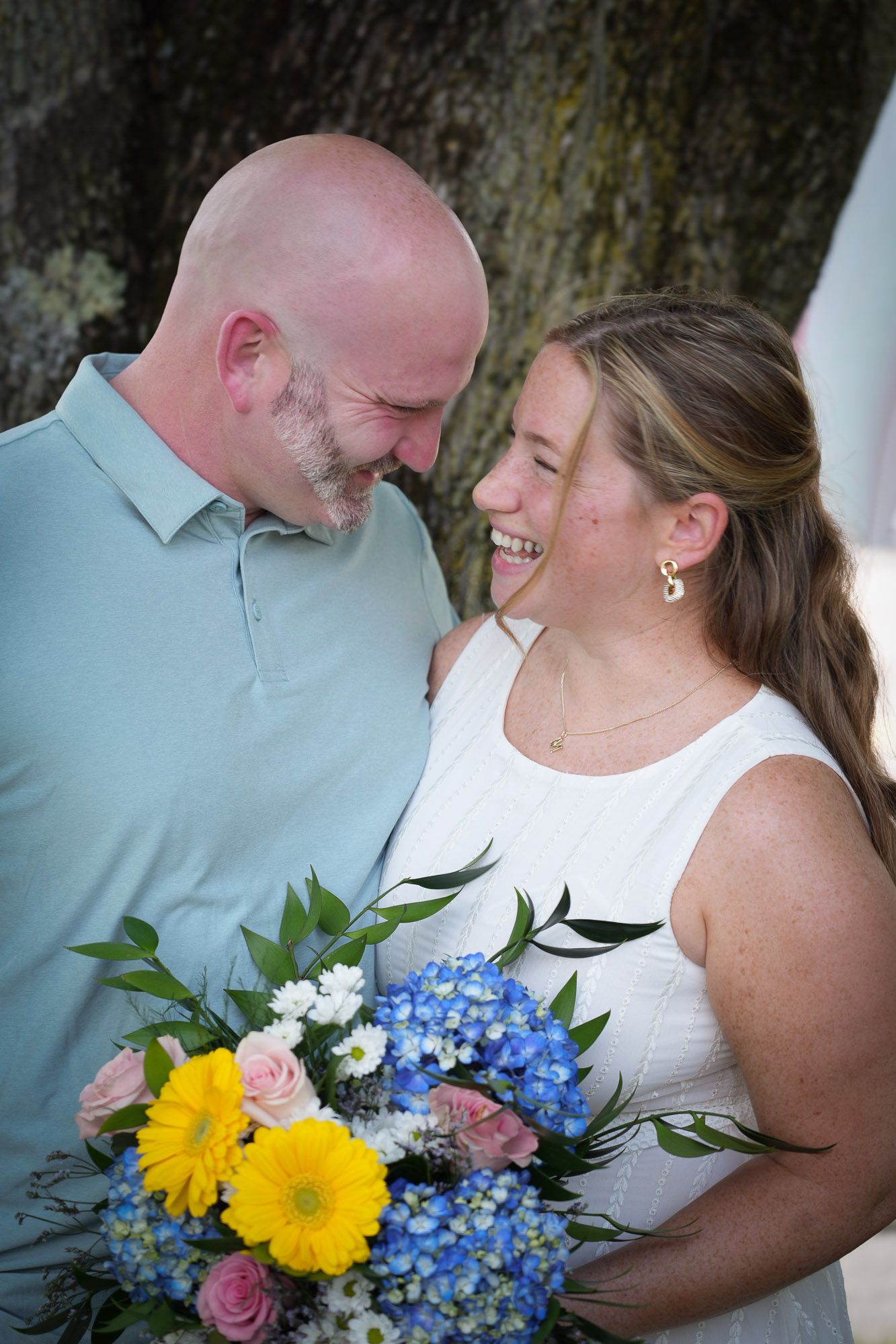 Happy newlywed couple laughing together under tree, bride holding colorful bouquet.