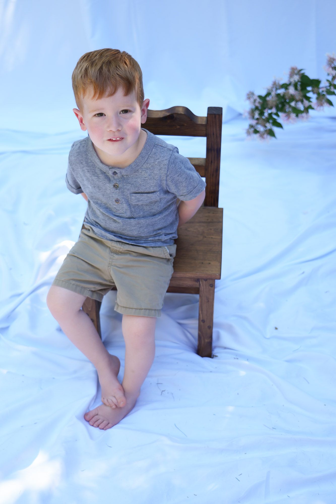 Cute redhead toddler boy sitting beside a wooden chair on white fabric backdrop.