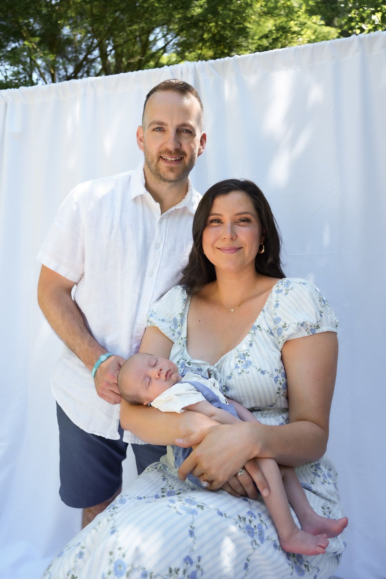 Family portrait of parents with newborn baby against white backdrop outdoors.