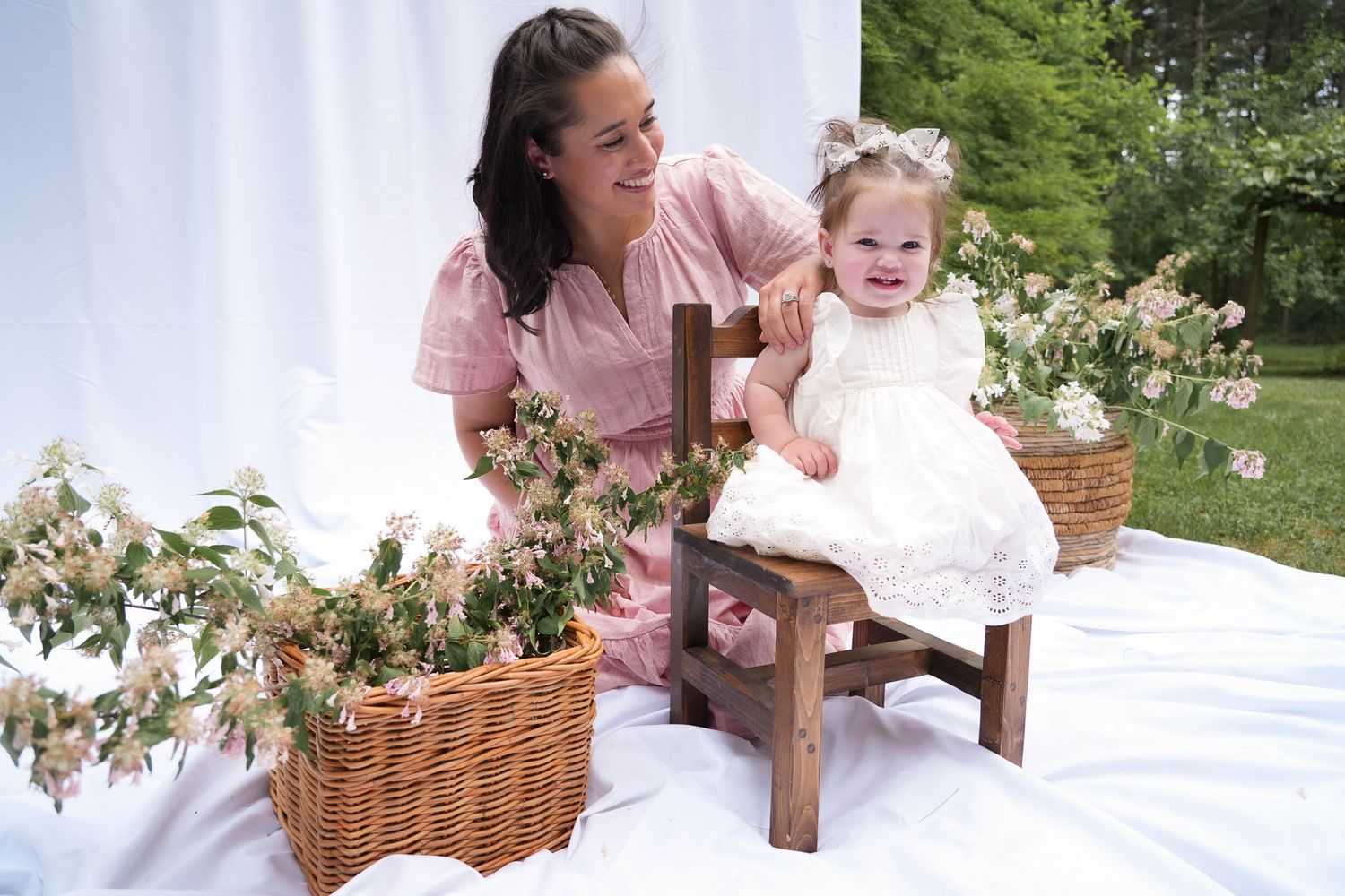 Mother in pink dress smiles at happy toddler girl in white lace dress seated on small wooden chair outdoors.