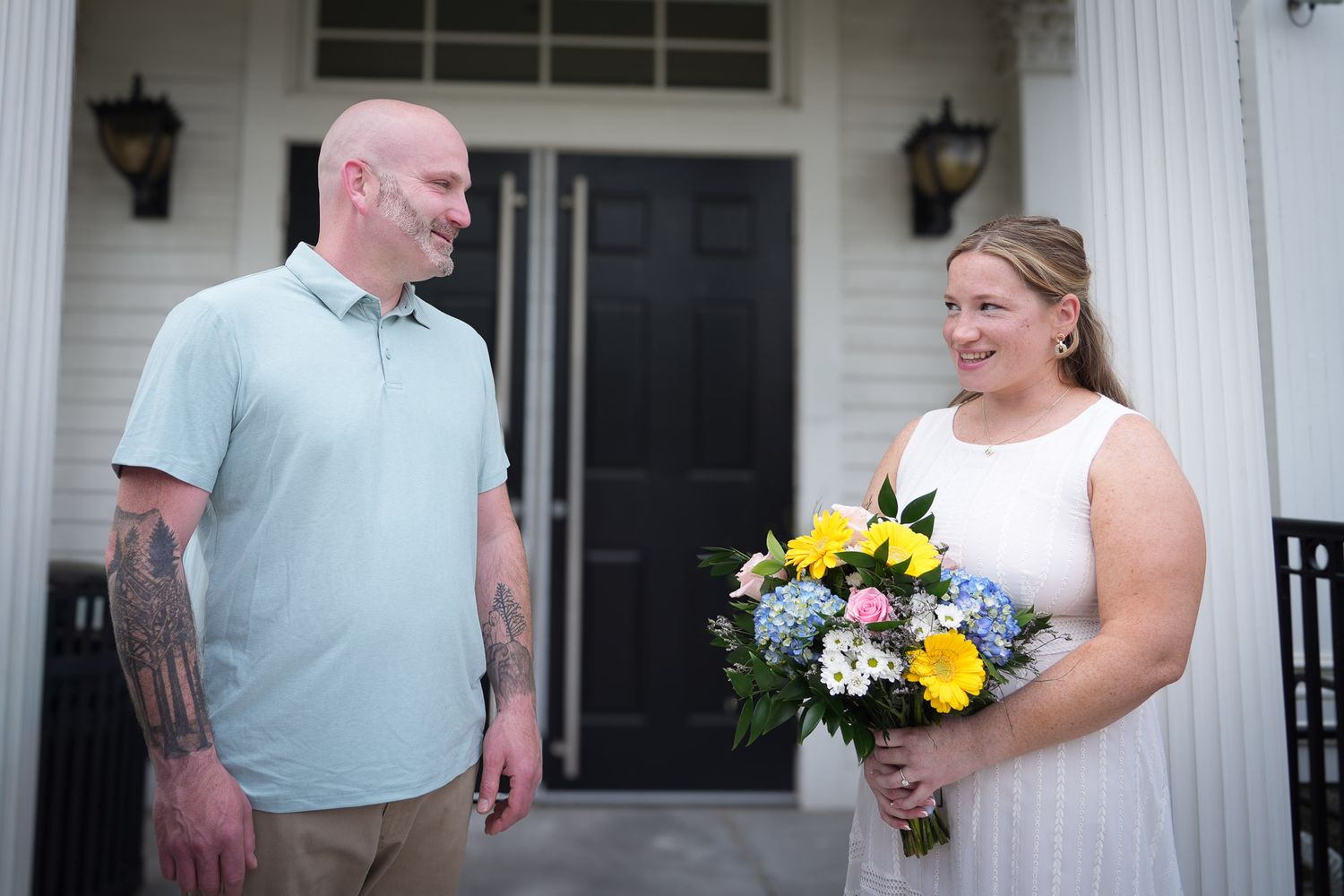 Couple sharing a joyful moment on porch steps, bride in white dress holding bouquet, groom in light blue polo.
