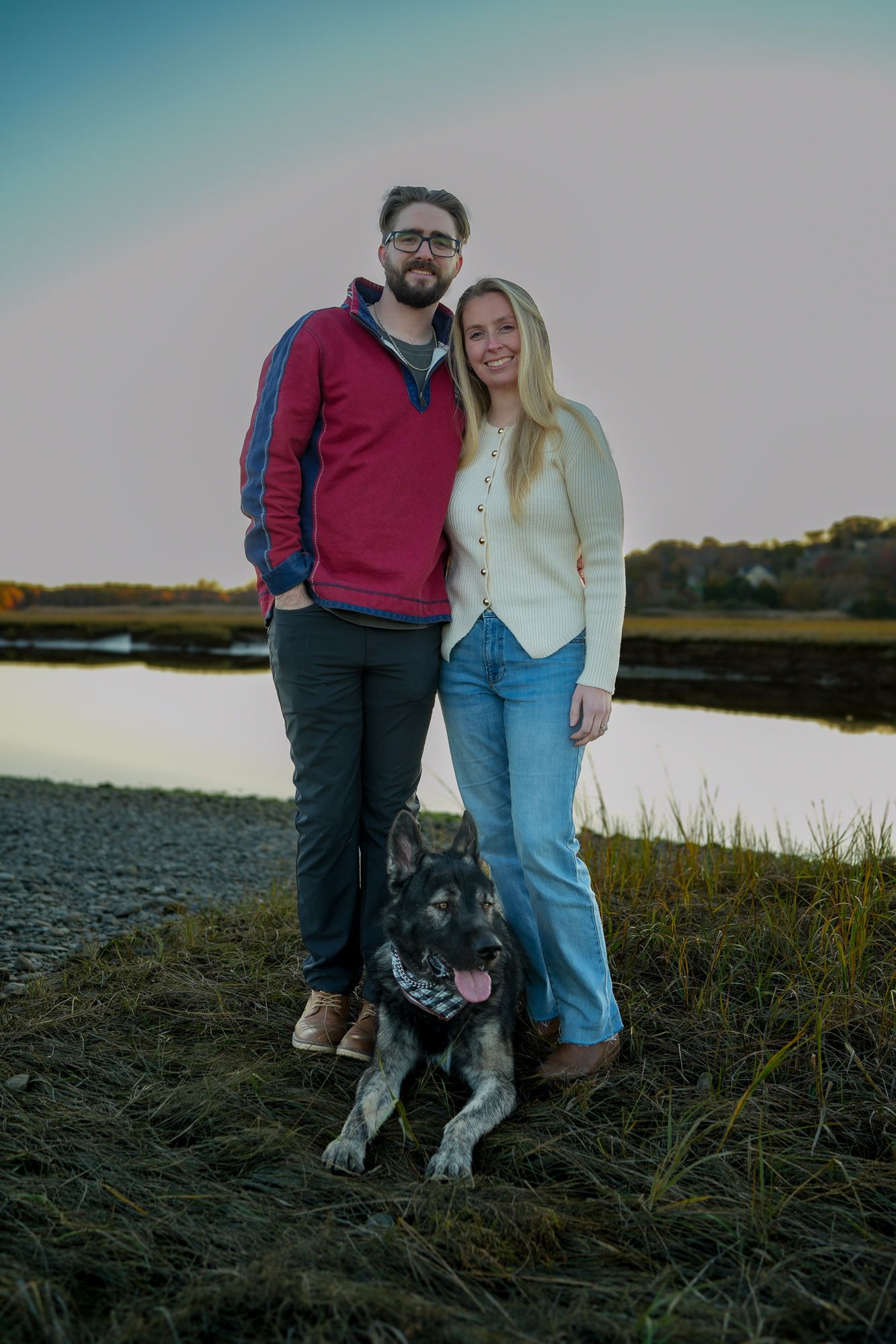 Couple standing with their German Shepherd dog by a calm marsh waterway at golden hour.
