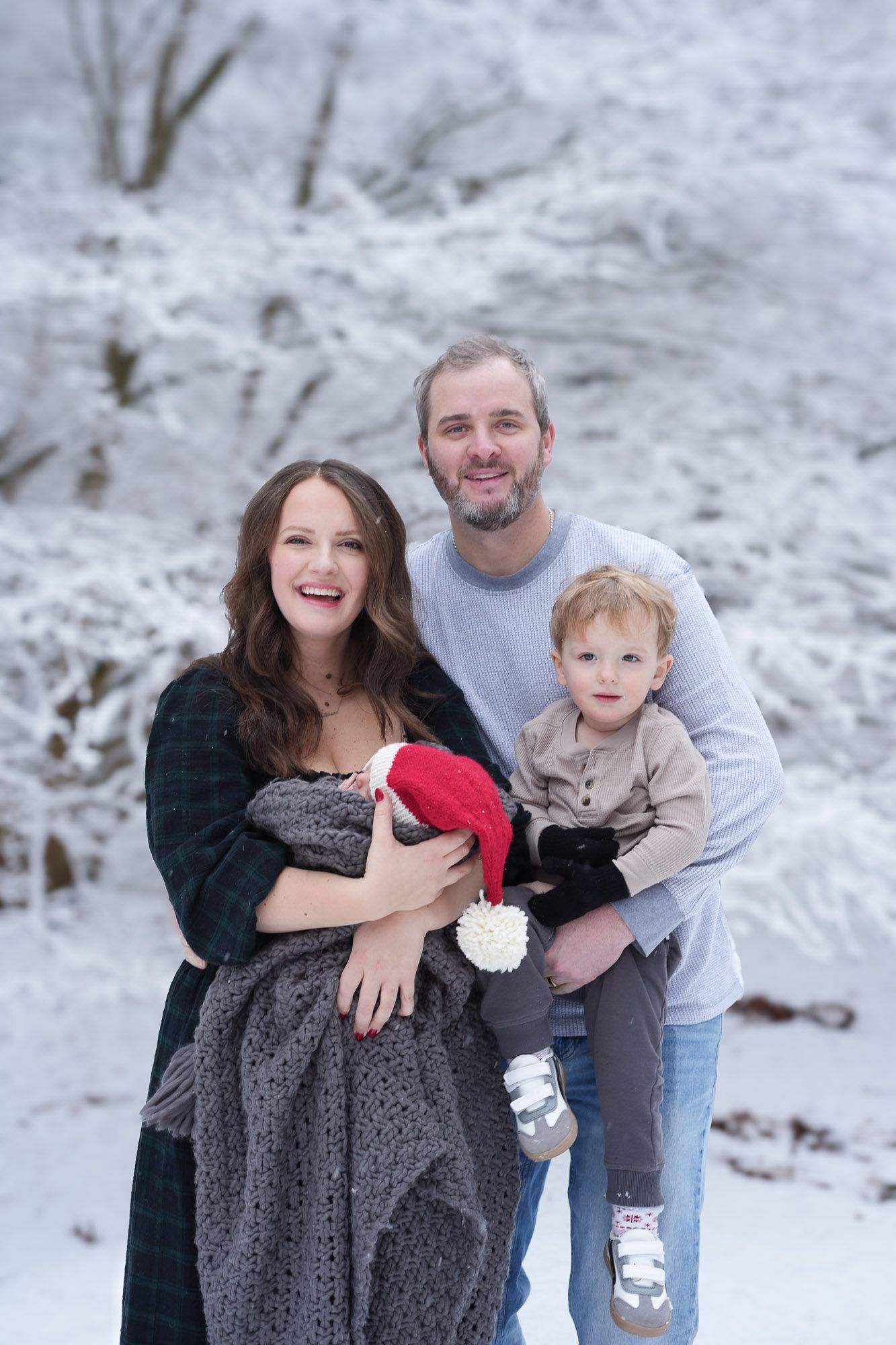 Happy family posing outdoors in snowy winter setting with toddler in Santa hat and newborn.