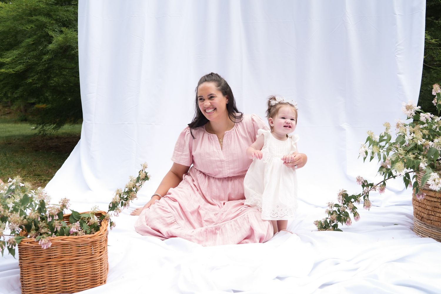 A mother in a pink dress sits on white fabric outdoors with her toddler daughter in a white dress beside flower baskets.