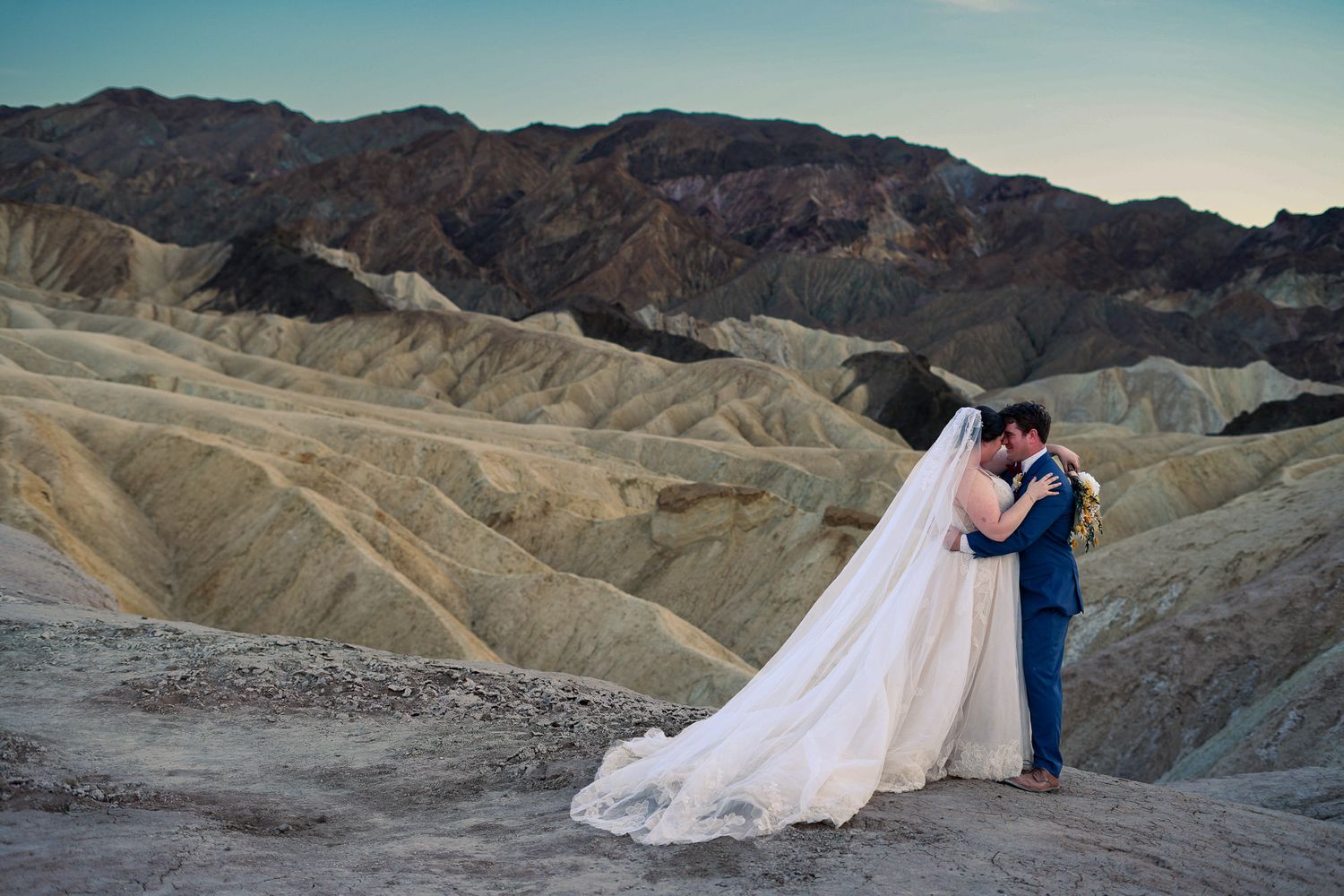 Newlyweds embrace at Zabriskie Point, bride in flowing veil gown, groom in blue suit at sunset.