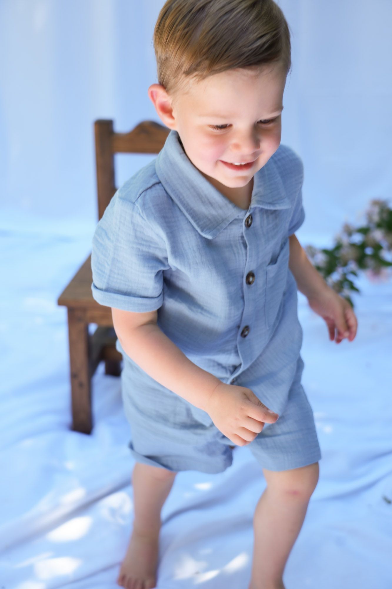Happy toddler boy in blue button-up outfit standing barefoot near wooden chair indoors.