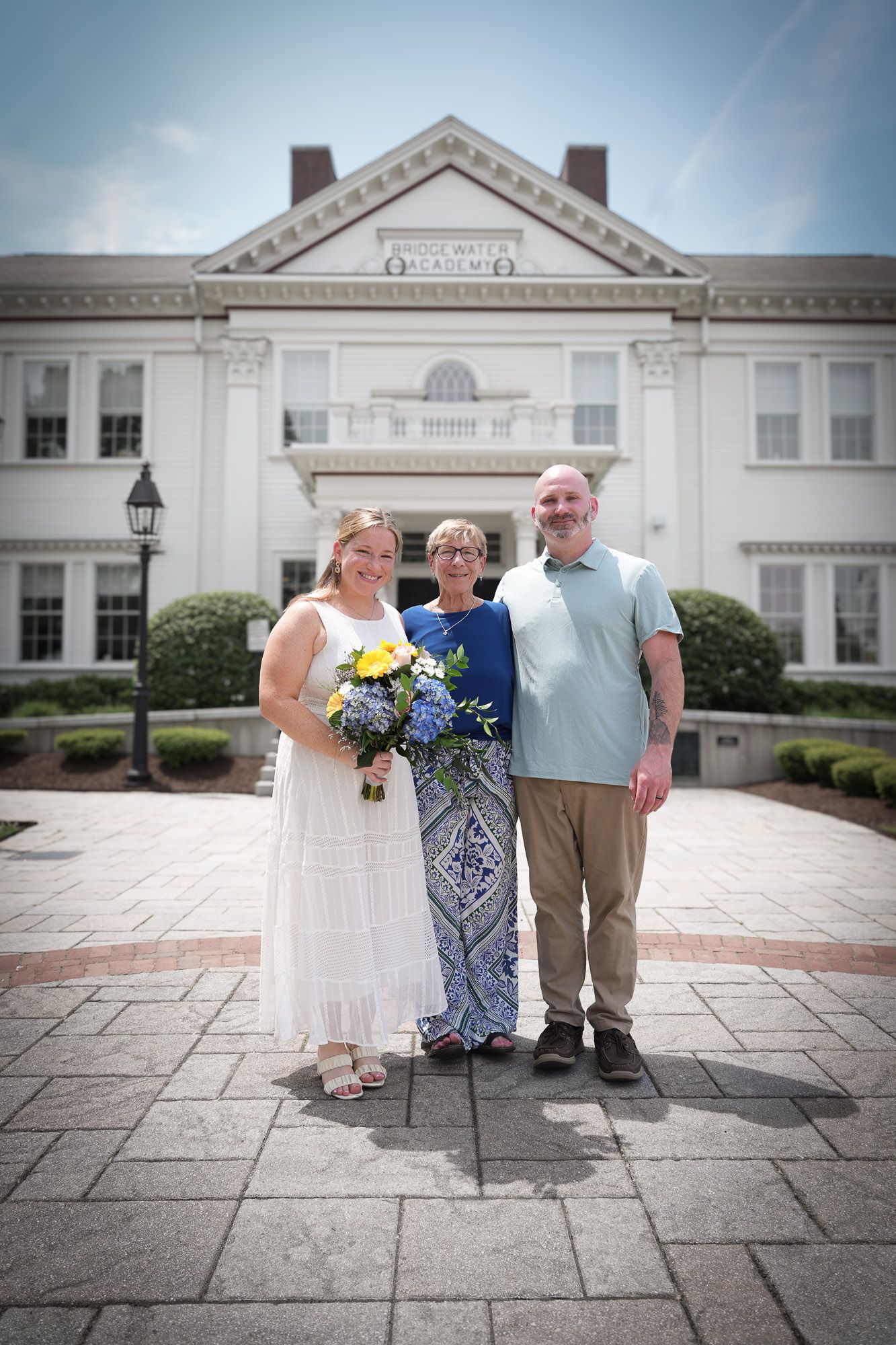 Wedding trio posing in front of Bridgewater Academy, bride holding yellow and blue bouquet.
