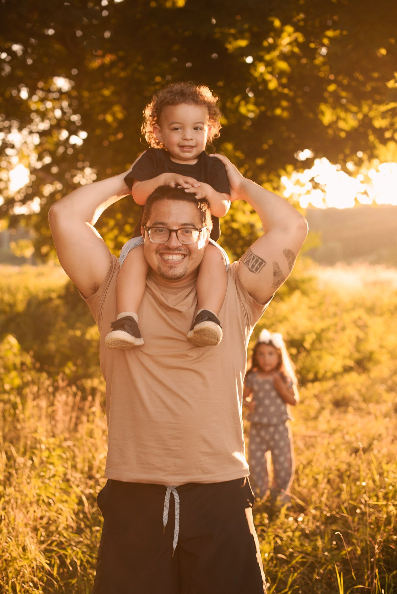 A smiling dad carries his toddler on his shoulders in a golden sunlit field with a child in background.