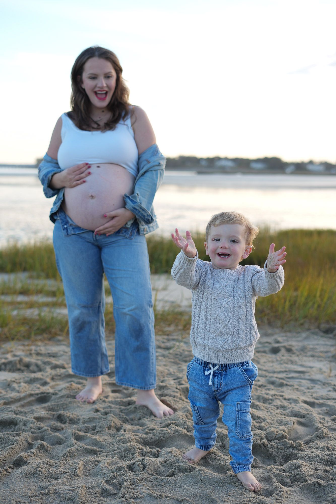 Excited toddler and pregnant mother in jeans posing together on beach at sunset.