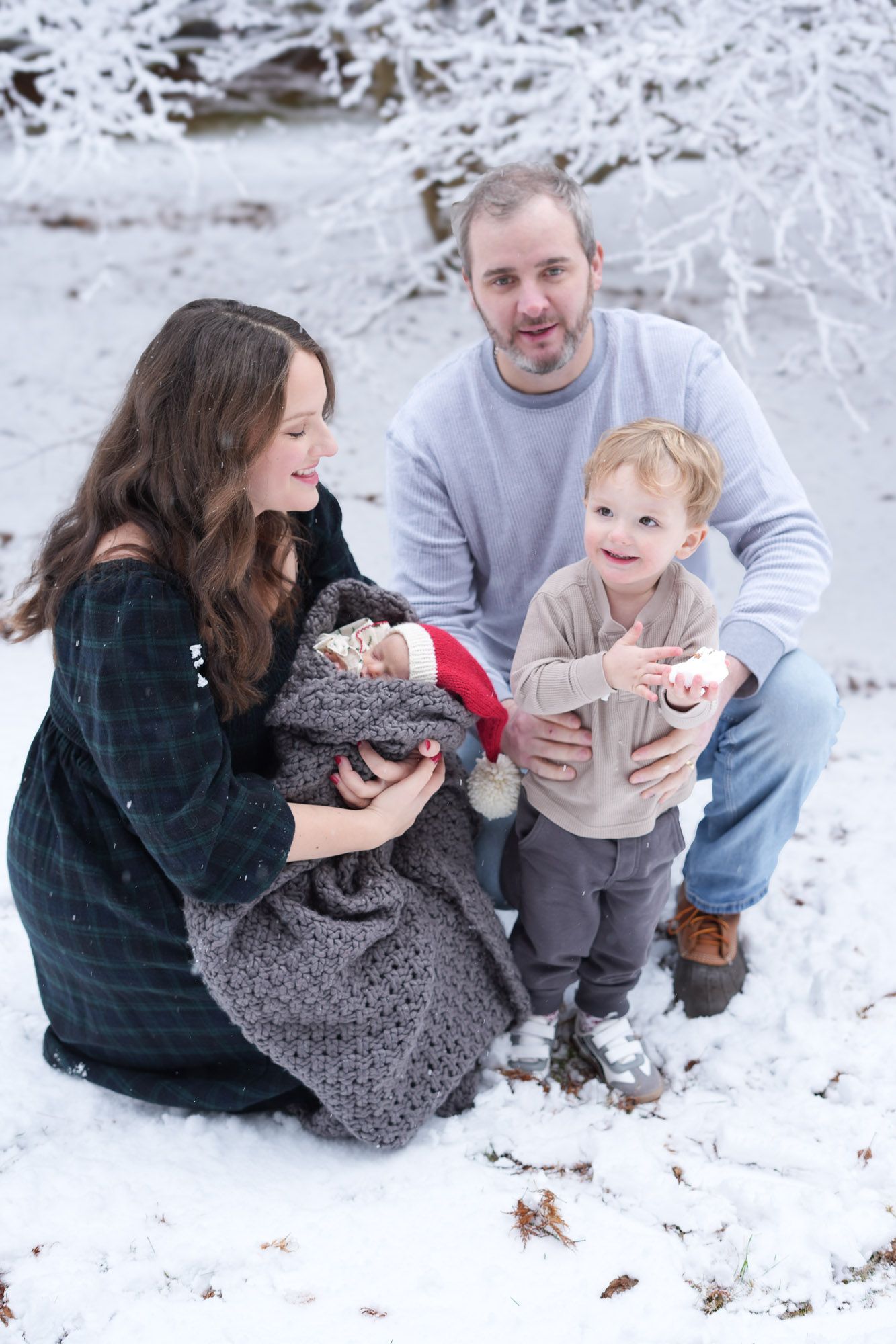 A joyful family poses in a snowy winter setting, with parents crouching beside their excited toddler son and newborn baby.