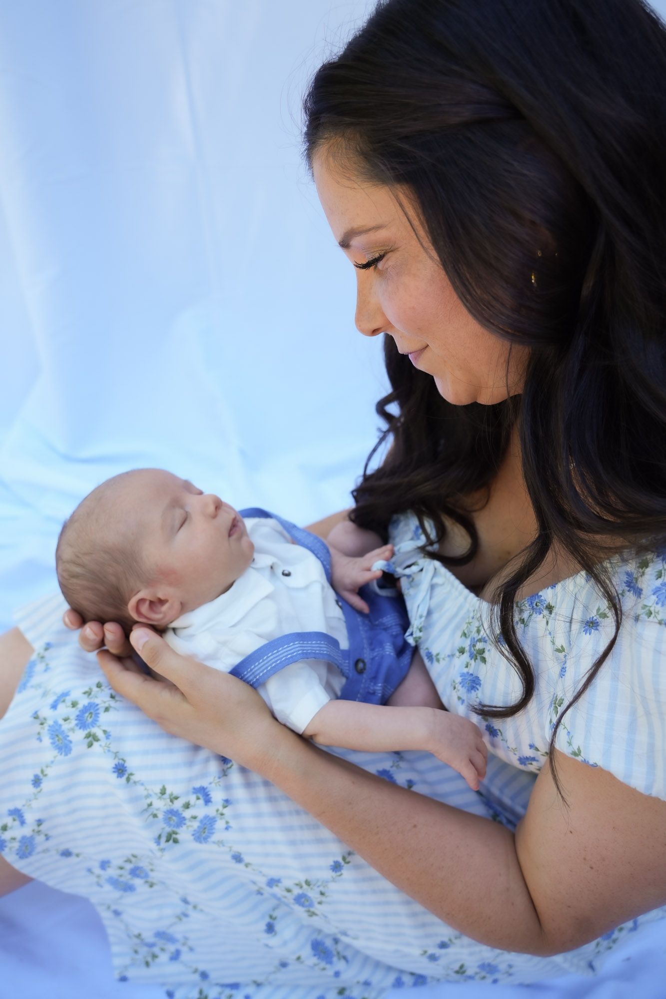 A mother with dark hair tenderly holds her newborn baby in a blue floral outfit against a soft blue backdrop.