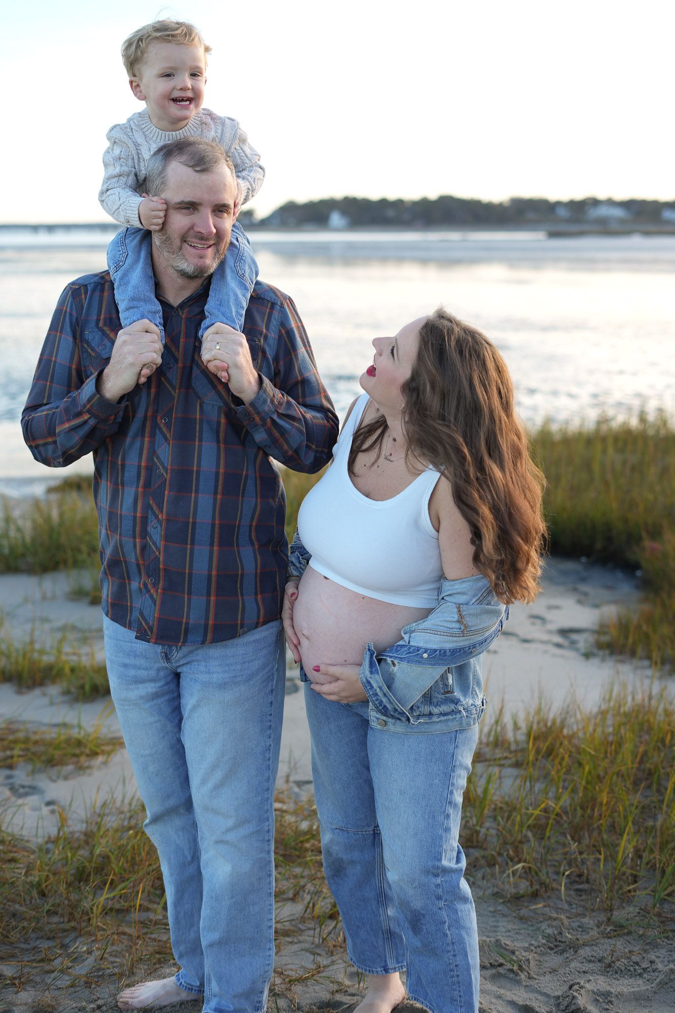 Pregnant woman and man with laughing toddler on shoulders sharing joyful moment on beach.