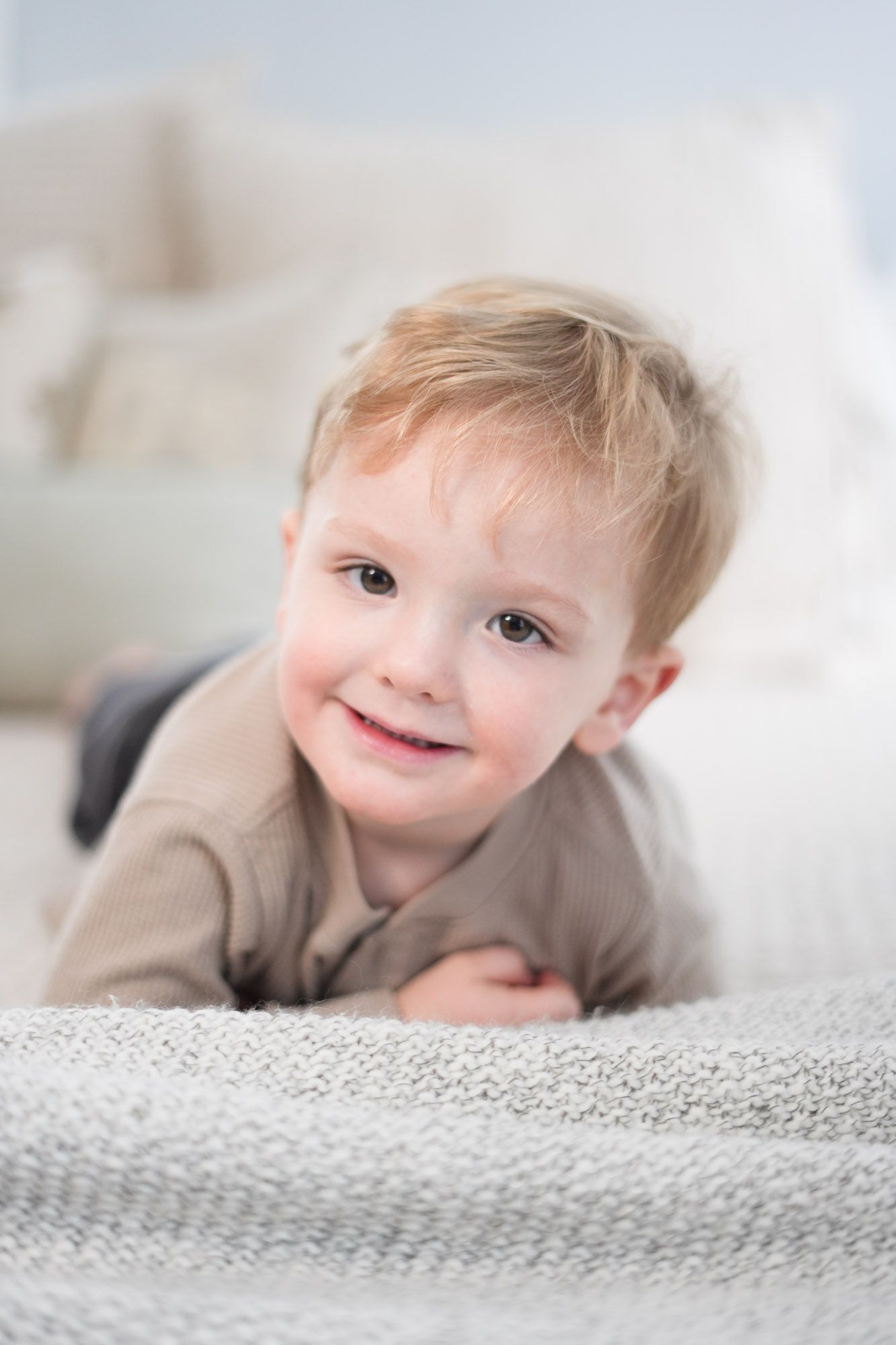 Smiling toddler boy with blonde hair lying on a gray knit blanket.
