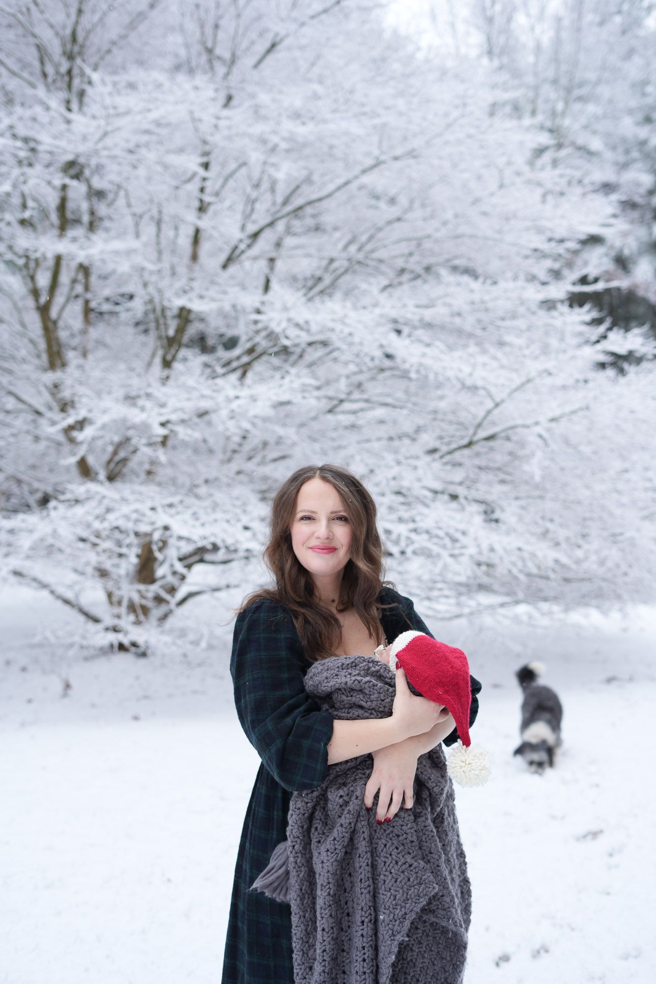 Smiling mother cradling newborn in gray blanket with Santa hat in snow-covered winter forest.